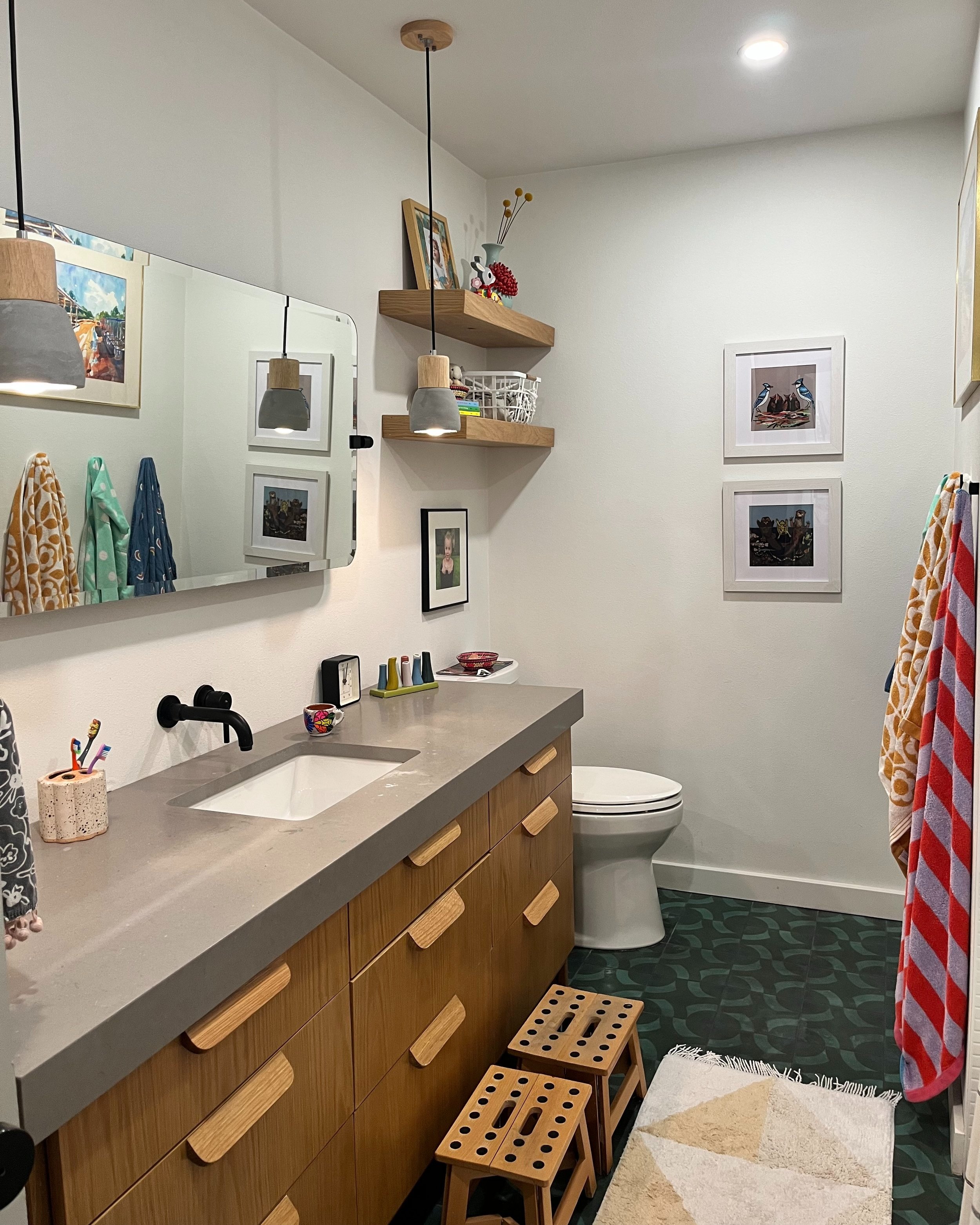 A bathroom with a large mirror, a gray countertop, and wooden cabinets. There are two small wooden stools in front of the cabinet, and a white toilet in the corner. The wall has framed pictures, and wooden shelves hold decorative items. Towels hang on a black rod, and a patterned rug is on the green tiled floor.