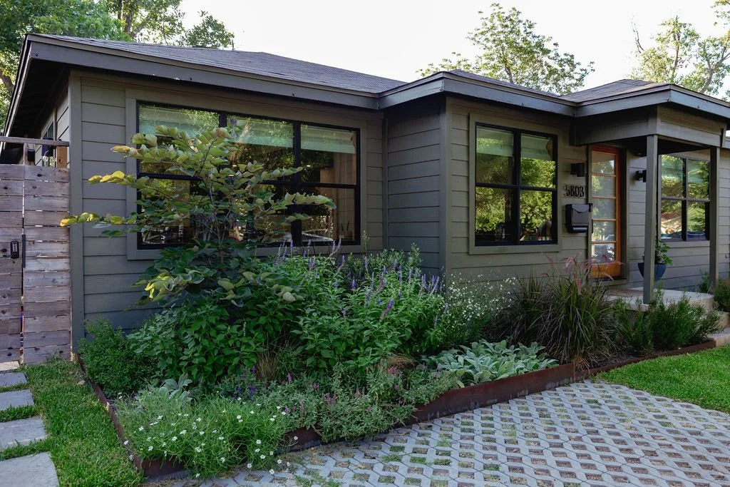 A modern gray house with a garden in front, featuring various green plants and purple flowers. The house has large black-framed windows and a small porch with a wooden door.