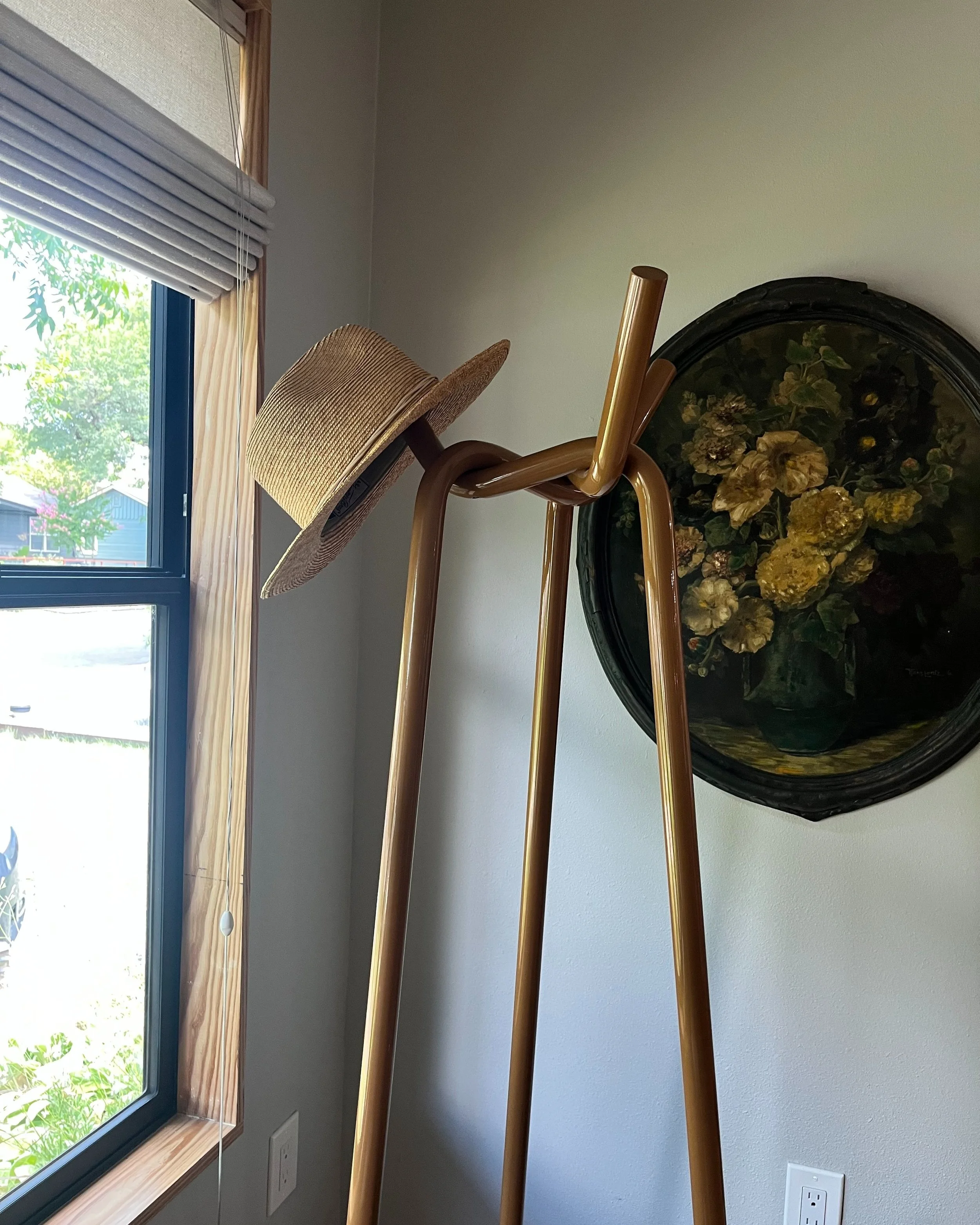 A corner of a room with a window, a straw hat resting on a wooden art stand, a decorative round framed floral painting on the wall, and electrical outlets below.