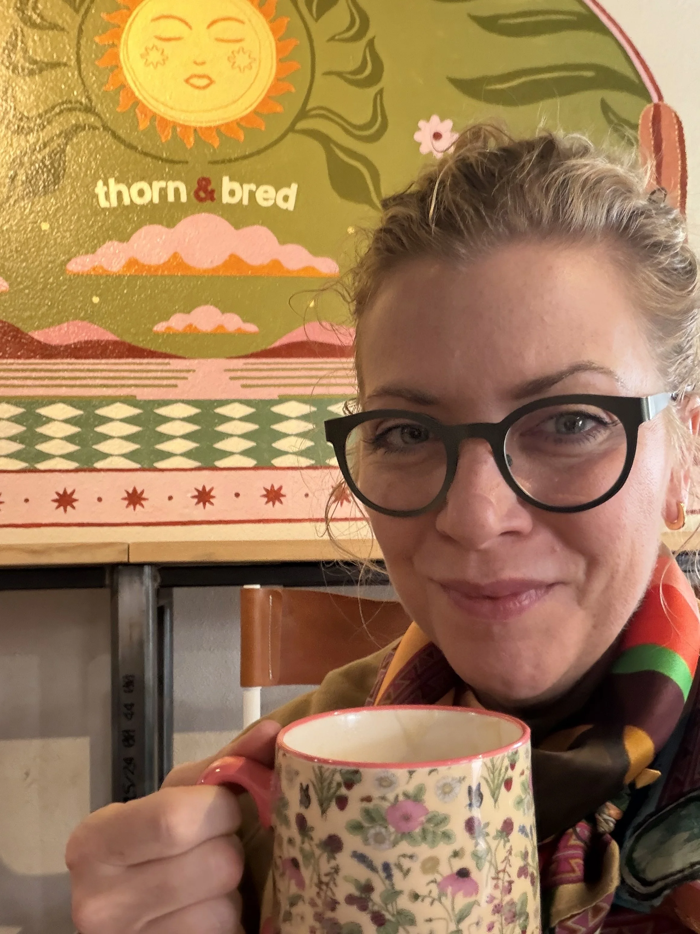 A woman with glasses holding a floral patterned mug, sitting indoors in front of a colorful wall art that reads 'thorn & bred' with a smiling sun illustration, sunflowers, and abstract design elements.