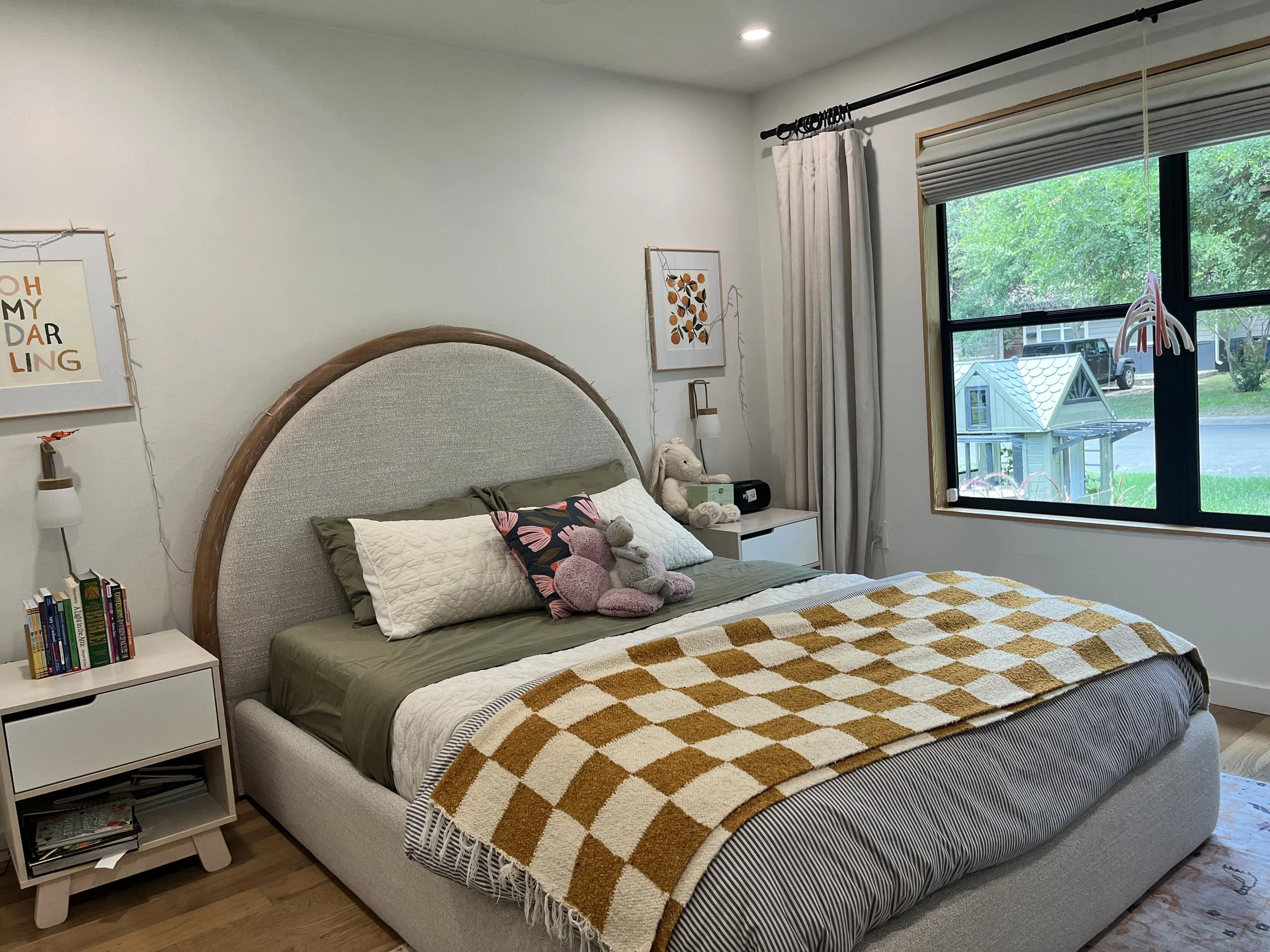 A cozy bedroom with a bed that has a beige upholstered headboard, a green bedsheet, and pillows with stuffed animals. There are two white nightstands, one on each side of the bed, with books and a lamp. The wall has framed art, one with colorful text and another with leaf illustrations. A large window with cream curtains shows a neighborhood scene outside with a playhouse and greenery.