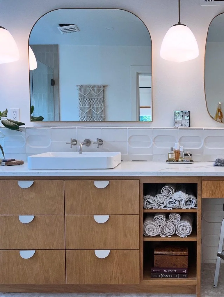 A modern bathroom vanity with a white rectangular sink, a large mirror, light wooden cabinets and drawers, and rolled white towels on a shelf. There are two hanging pendant lights and a bottle of soap or lotion on a tray.