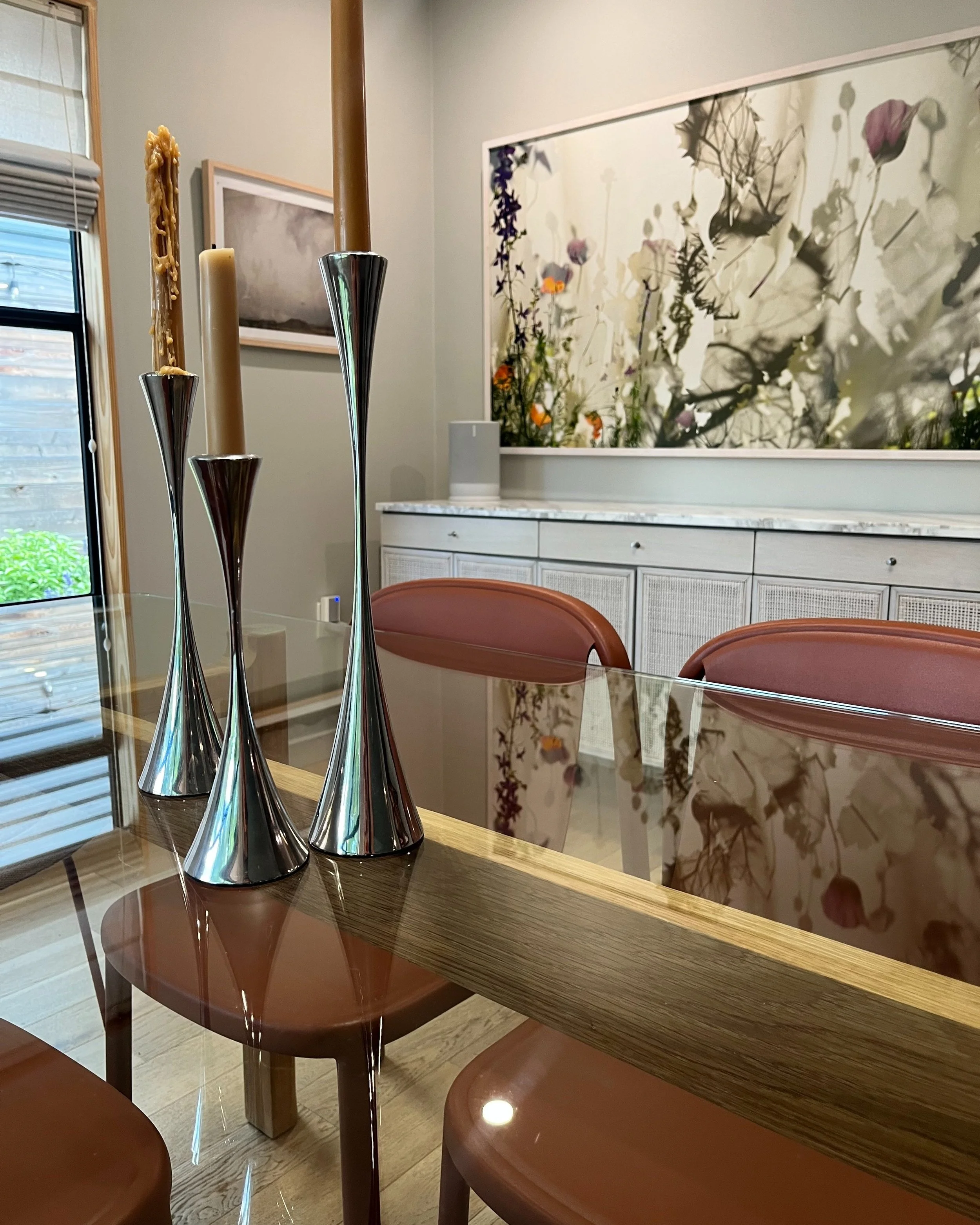 A dining room with a glass table, pink chairs, and a multi-colored abstract floral art piece on the wall. Tall silver candlesticks with candles are placed on the table.