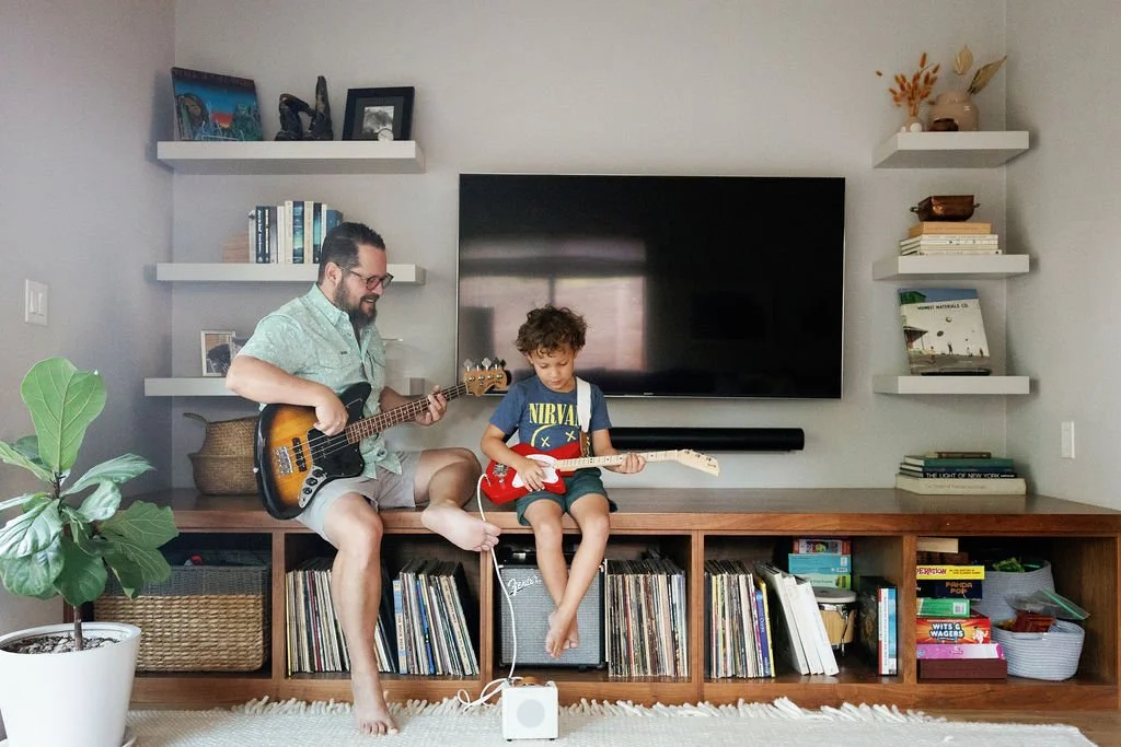 A man and a young boy sitting on a wooden entertainment center, playing electric guitars together in a living room. The man is smiling, wearing glasses and a light-colored shirt, while the boy is focused, wearing a Nirvana t-shirt. There are books, records, and shelves with artwork and decor around them, with a large television on the wall behind them.