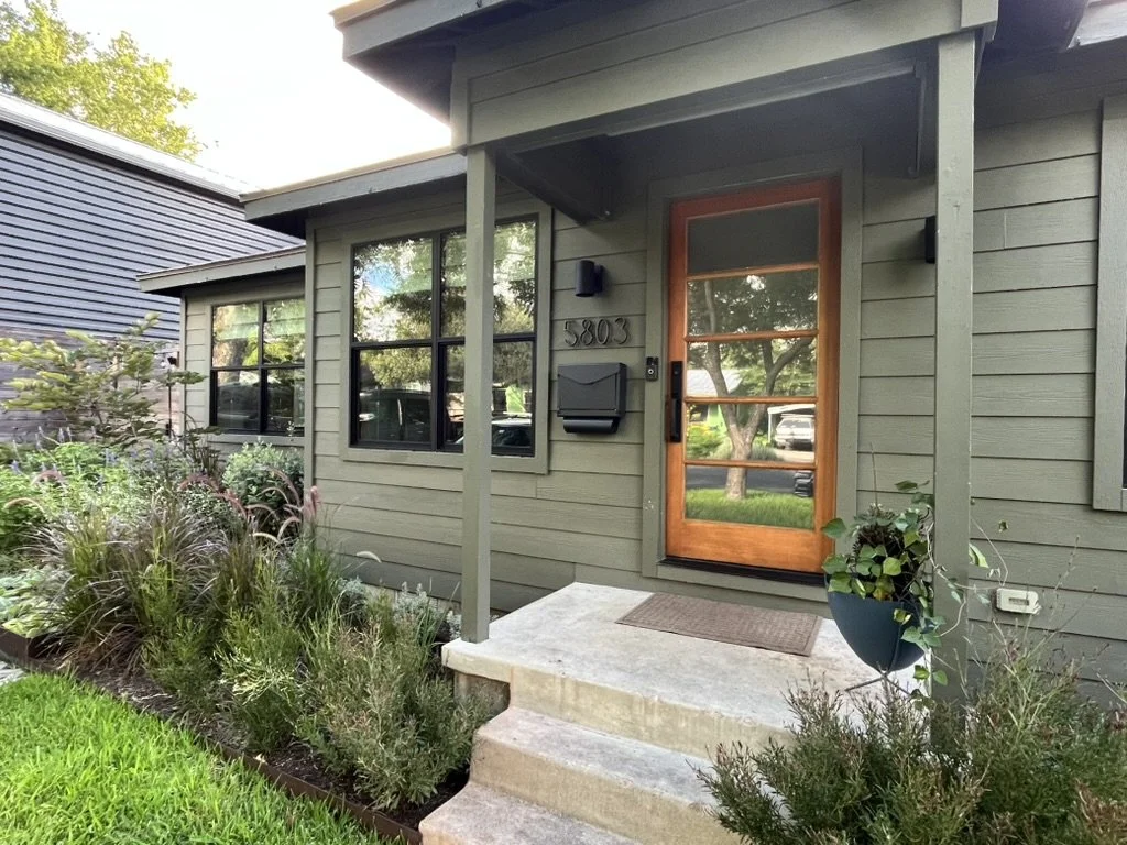 Front door of a house with a glass-paneled wooden door, surrounded by green shrubbery and plants, with steps leading up to the entrance and a black mailbox beside the door.