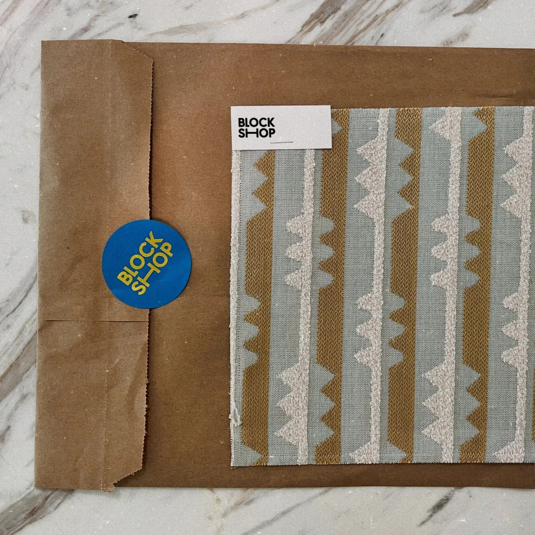 Fabric sample with geometric pattern in beige, light blue, and white, labeled BLOCK SHOP, attached inside a brown paper folder with BLOCK SHOP sticker, placed on a marble surface.