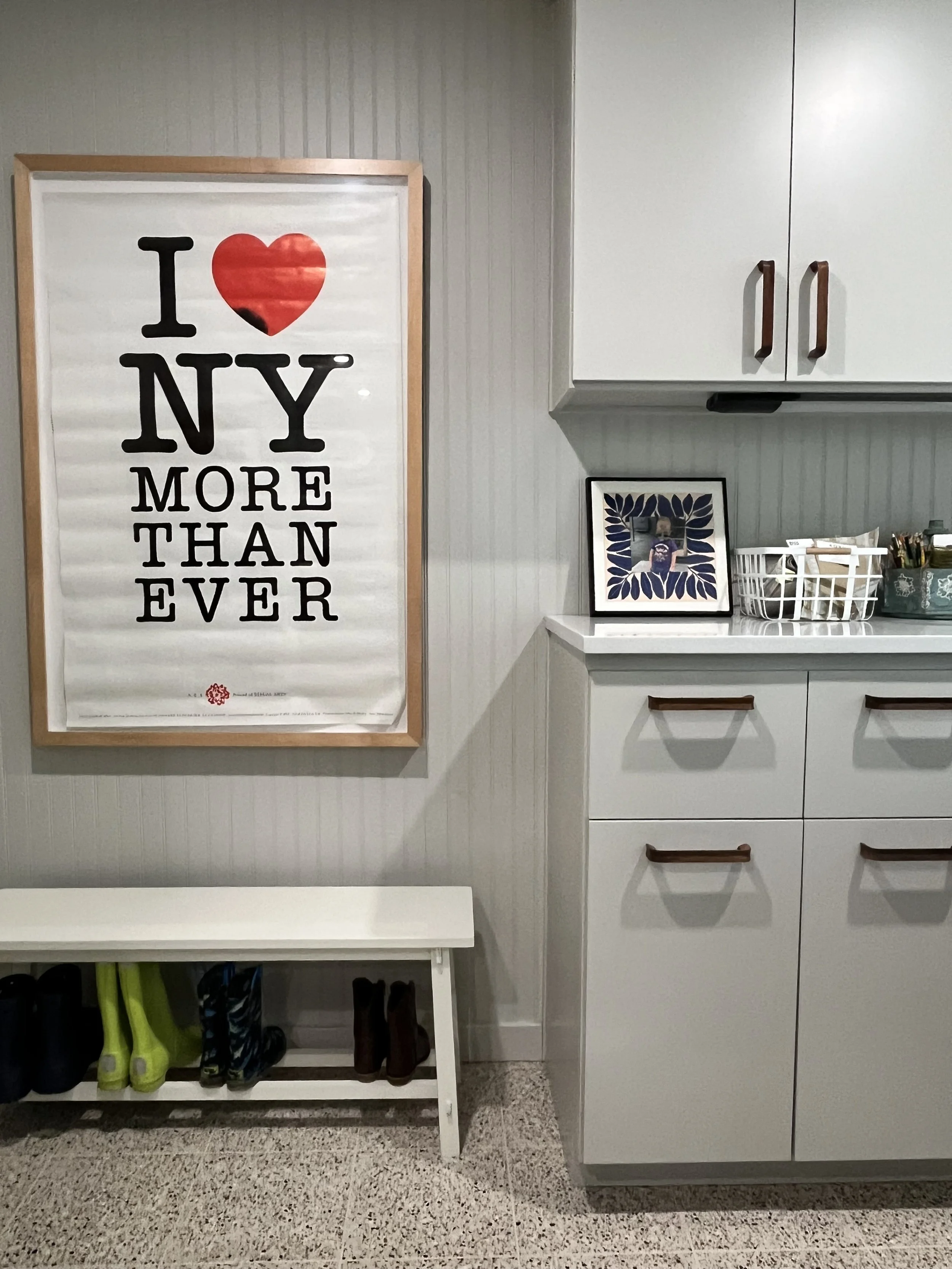 A framed poster on a wall with the text "I ♥ NY MORE THAN EVER". Below the poster is a white bench with shoes stored underneath. To the right, there is a white cabinet with a framed photo, a basket of miscellaneous items, and some small containers on the countertop.