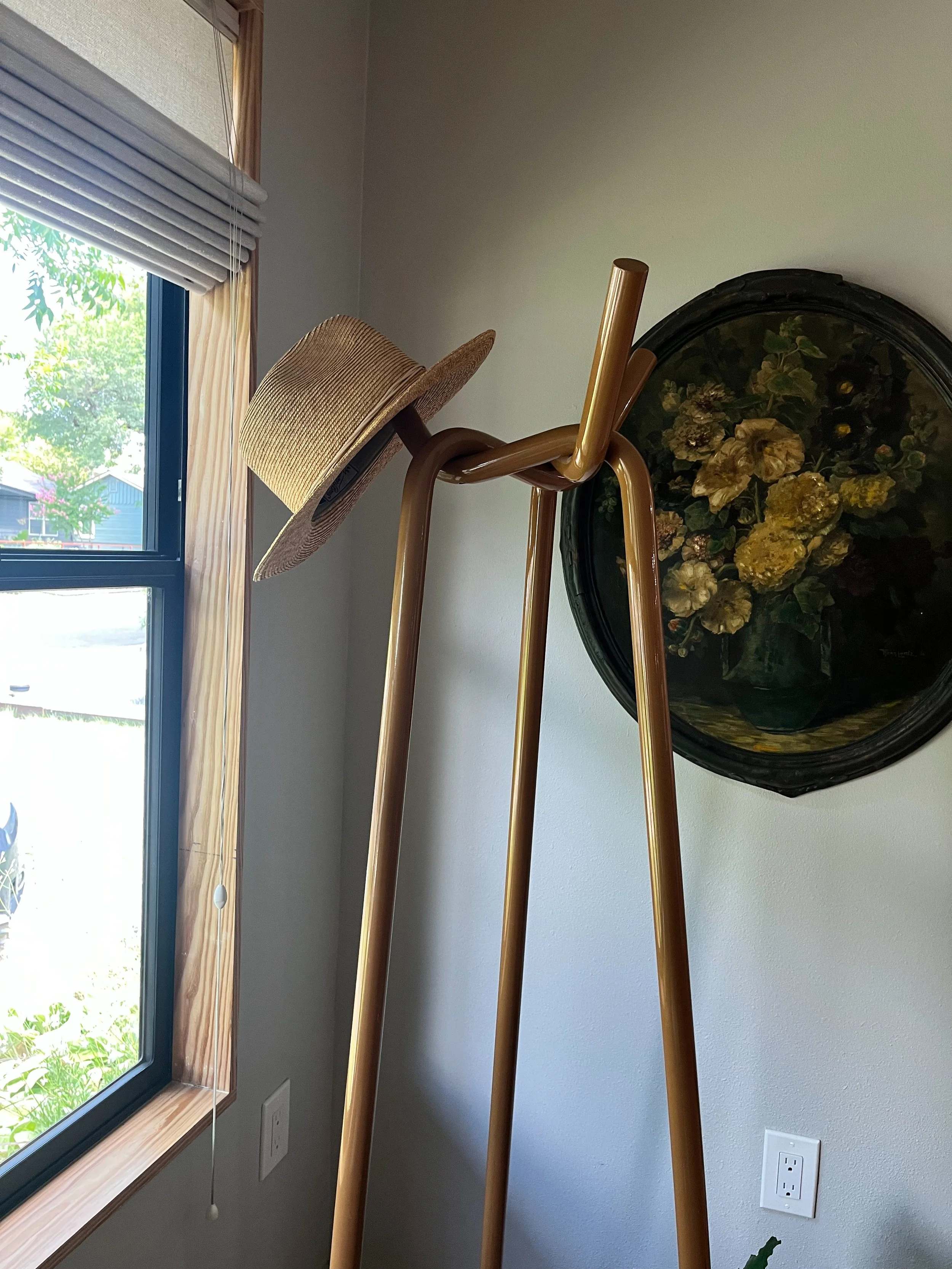 A corner of a room with a floor lamp that has a hat hanging on its top. There is a window with wooden trim on the left, and a wall with a round framed floral painting or mirror.