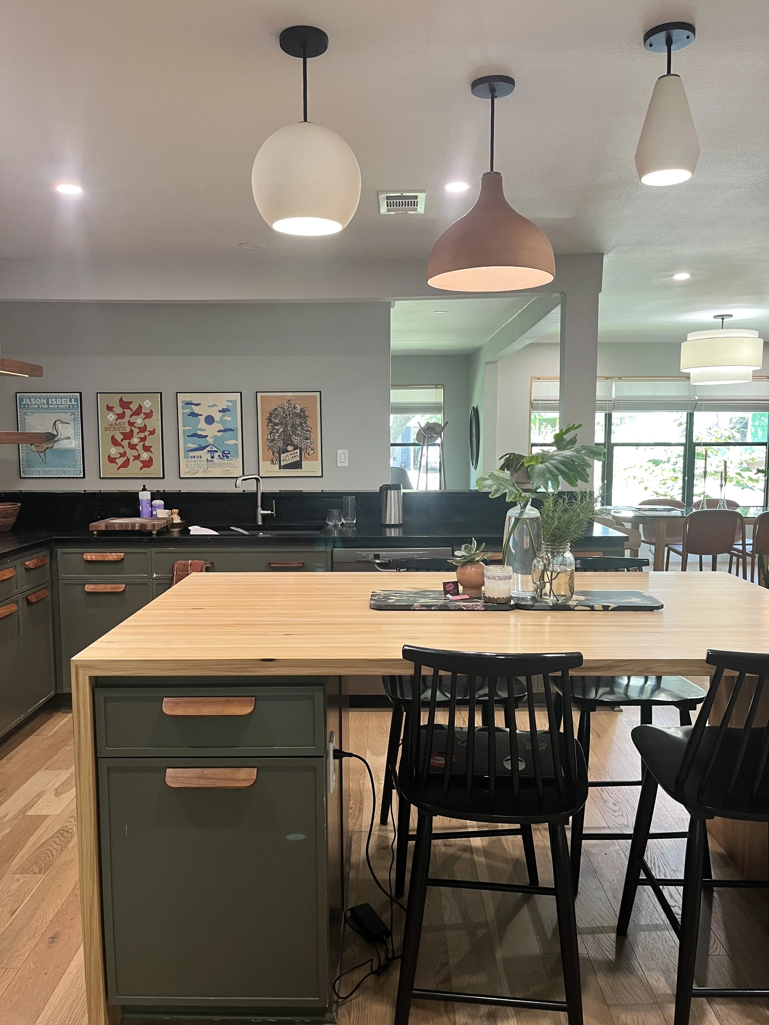 Modern open-concept kitchen and dining area with a large wooden island, black chairs, pendant lighting, green cabinets, and wall art, with sunlight through large windows.