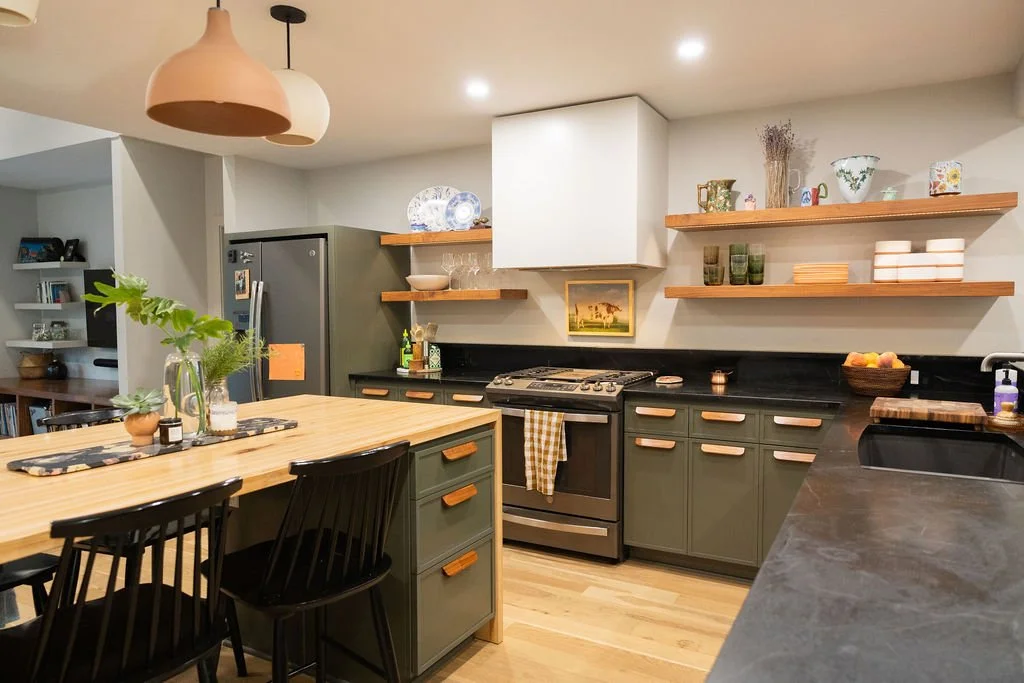 Kitchen with dark green cabinets, blonde wood countertops, open wooden shelves with dishes and decorative items, stainless steel refrigerator, black stove, and a wooden dining table with black chairs.