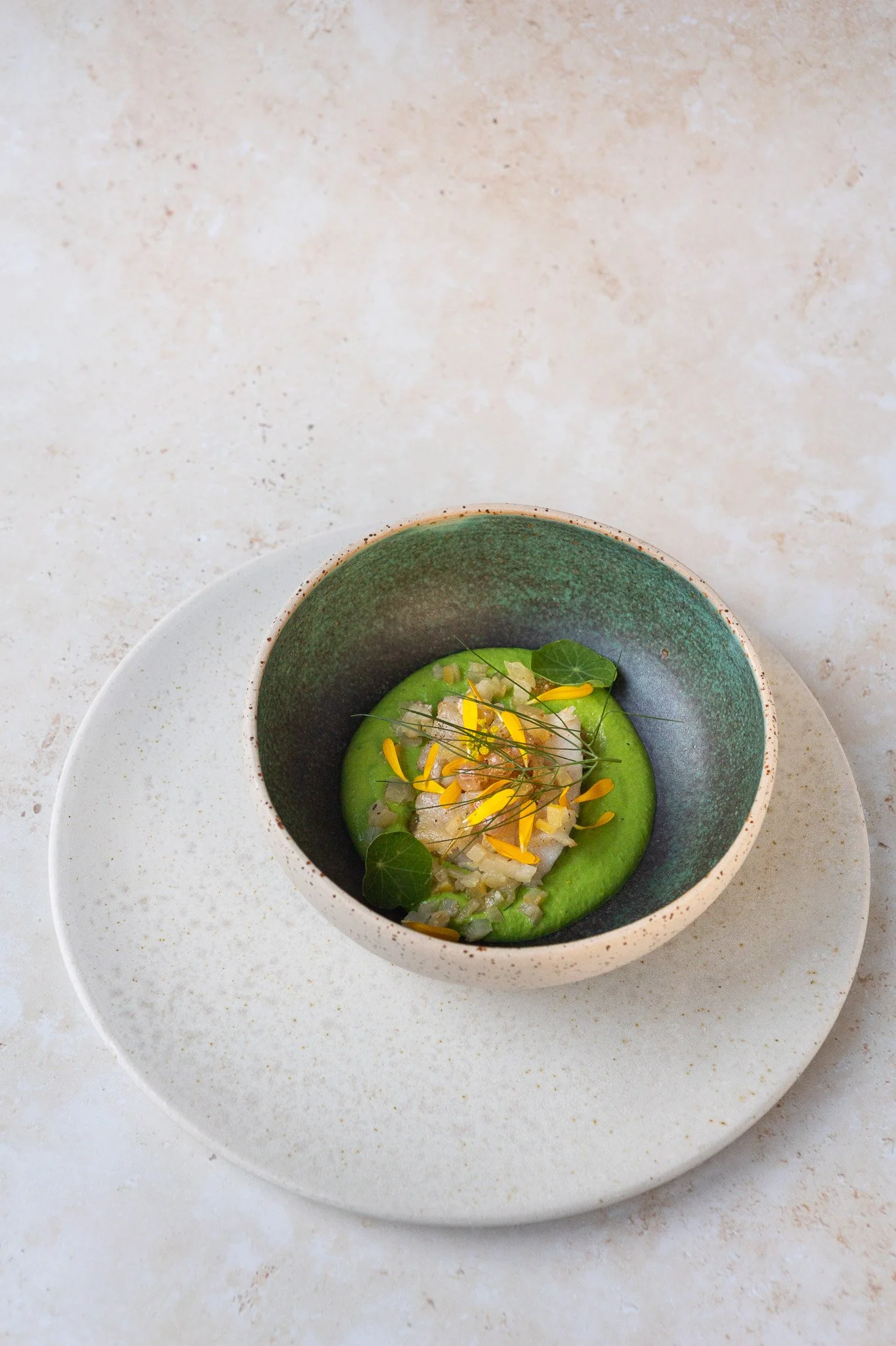 Ein Teller mit einer grünen cremigen Suppe, garniert mit gelben Blütenblättern, kleinen grünen Blättern und gehackten Zutaten auf einem weißen, runden Teller.