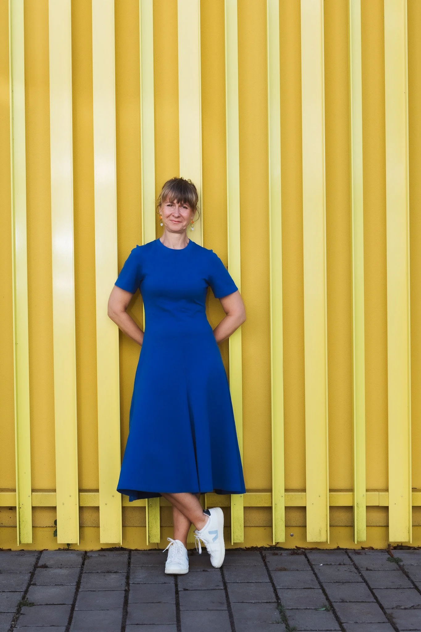 Frau in blauer Kleid steht vor einer gelben Wand mit vertikalen Strukturen, trägt weiße Sneakers, hat braune Haare mit Pony.