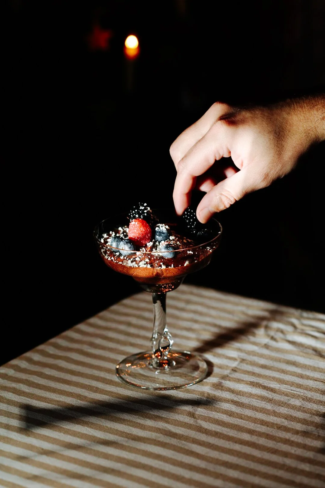Eine Hand garniert ein Glas mit Schokoladen-Dessert und Beeren, darunter eine braune und weiße Streifen-Tischdecke, im Hintergrund eine Kerze.