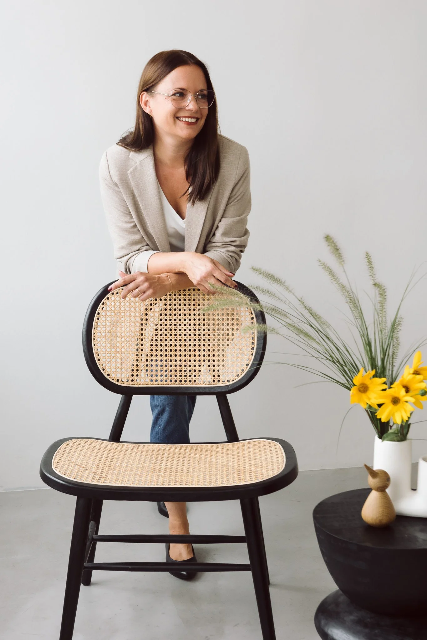 Frau in beige Blazer posiert hinter einem Stuhl mit Rattan-Sitzfläche, neben einem schwarzen Tisch mit Vase voller Sonnenblumen und grünen Zweigen.