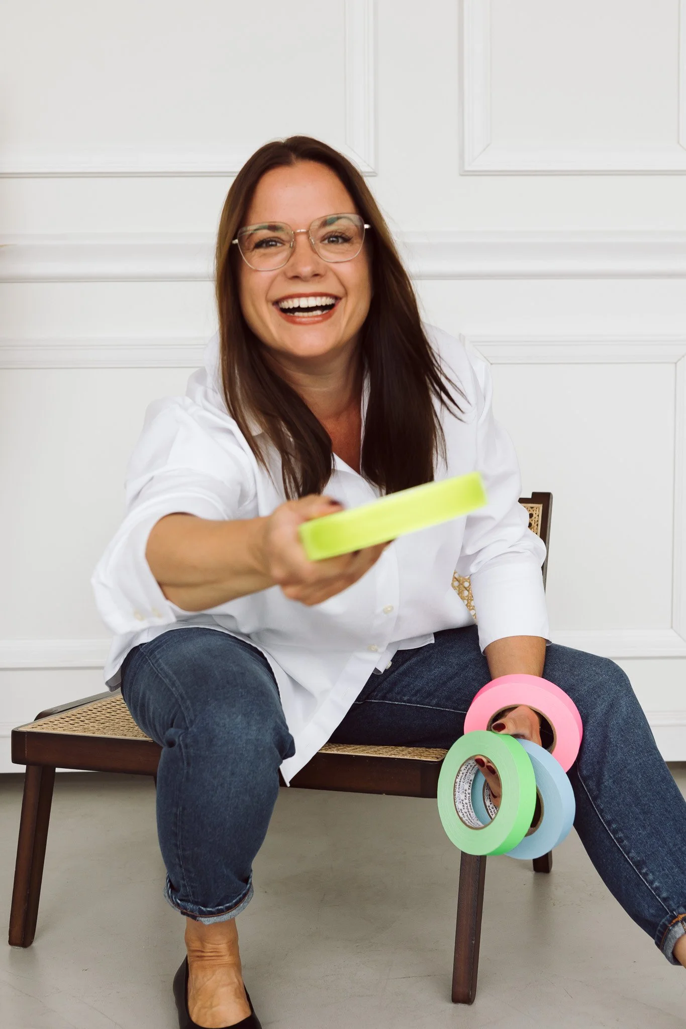Lächelnde Frau sitzt auf einem Stuhl, hält buntes Klebeband in der Hand und hat noch mehrere Rollen Klebeband in der Nähe.