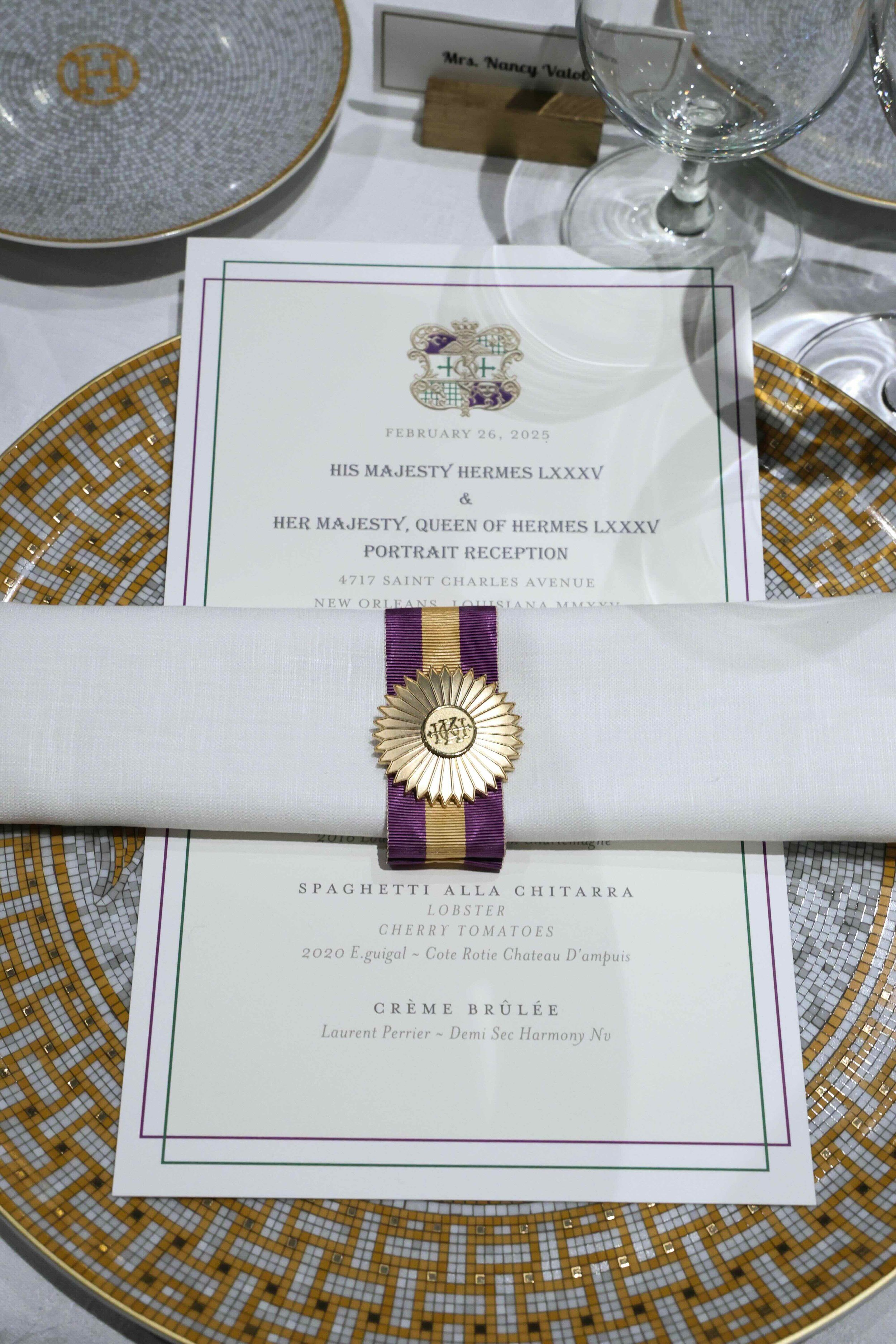 Formal table setting with a menu, a decorated napkin with a medal, two wine glasses, and patterned plates, indicating a special event or celebration.