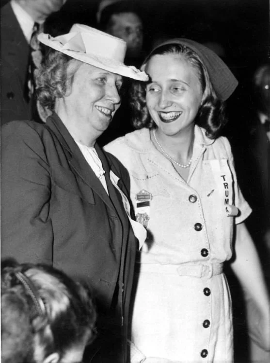 Two women smiling and talking at a gathering, one wearing a hat and the other with a 'Trump' campaign button on her dress.