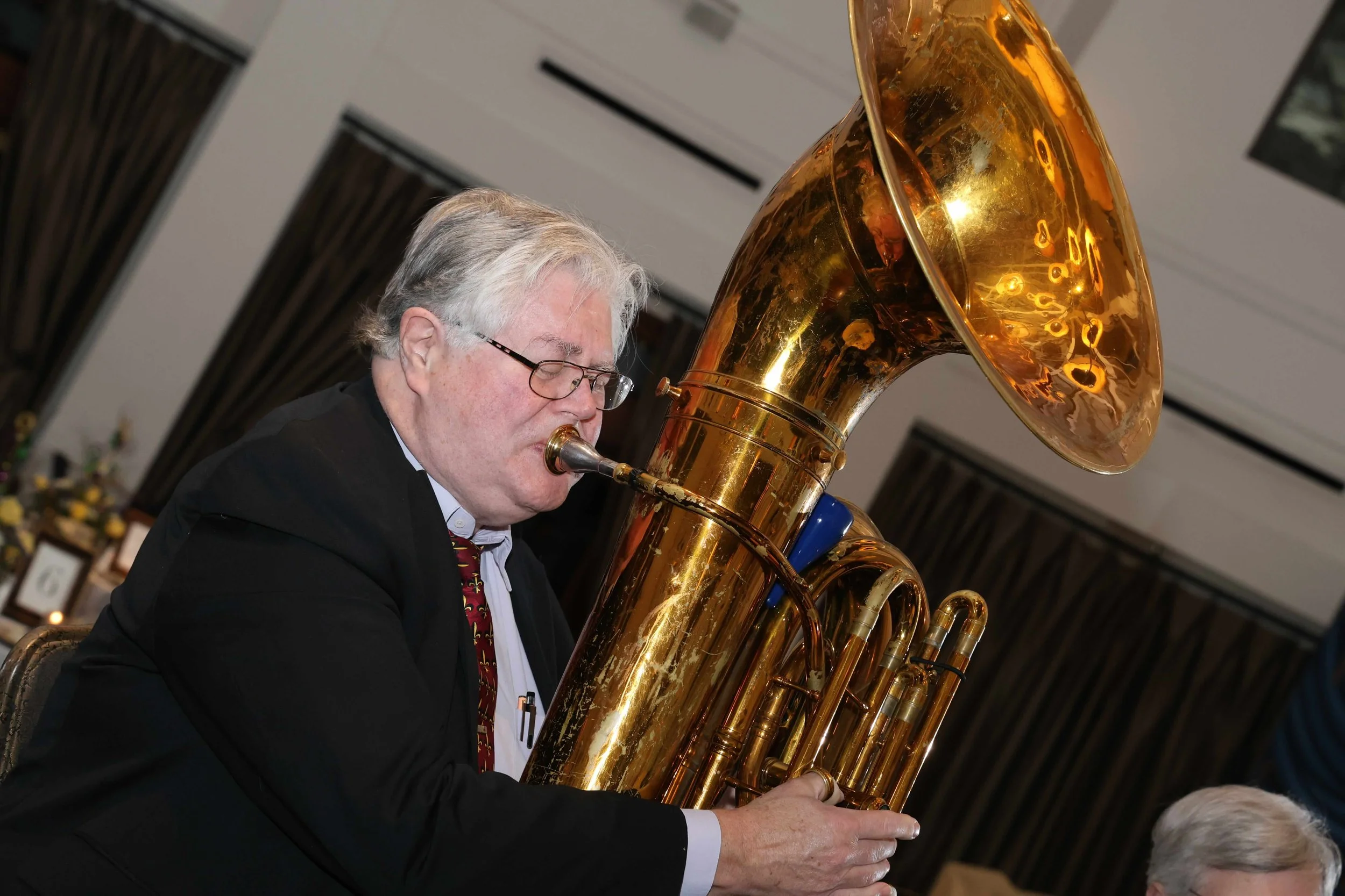 A man with gray hair and glasses playing a large brass tuba at an indoor event.