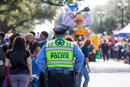 A New Orleans police officer in a neon green vest and blue uniform standing among a crowd at a parade or festival, with a large colorful float and trees in the background.