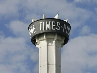 A water tower with a circular top displaying the text 'ONE TIMES-PIC', against a cloudy sky.