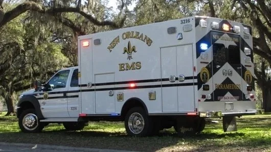 New Orleans EMS ambulance parked on grass under trees.