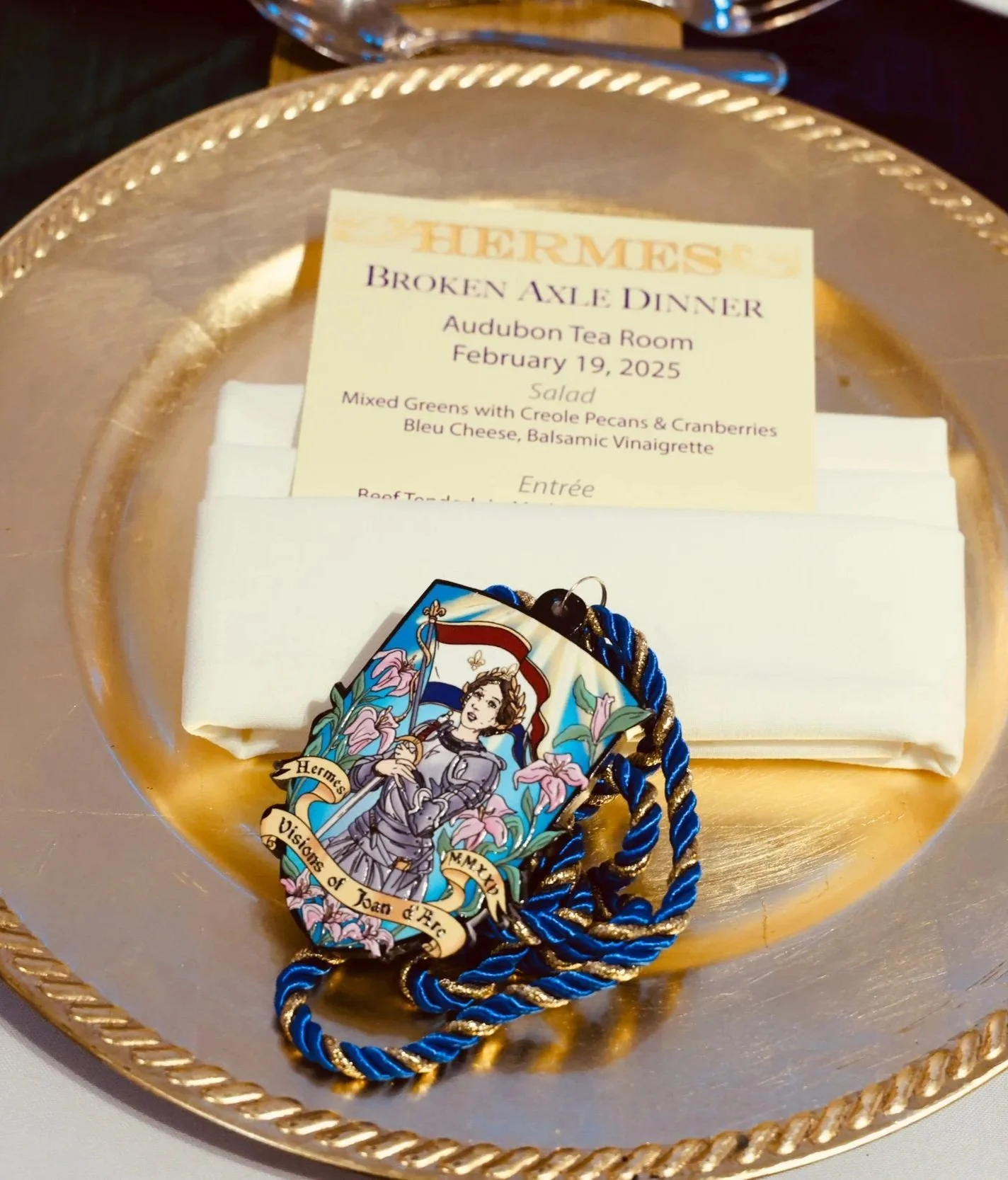 A golden tray holding a menu, a white cloth, a badge, and a blue and gold braided ribbon. The menu is titled 'Broken Ankle Dinner' from the Audubon Tea Room, dated February 19, 2025, featuring a salad of mixed greens with creole pecans and cranberries, bleu cheese, and balsamic vinaigrette.