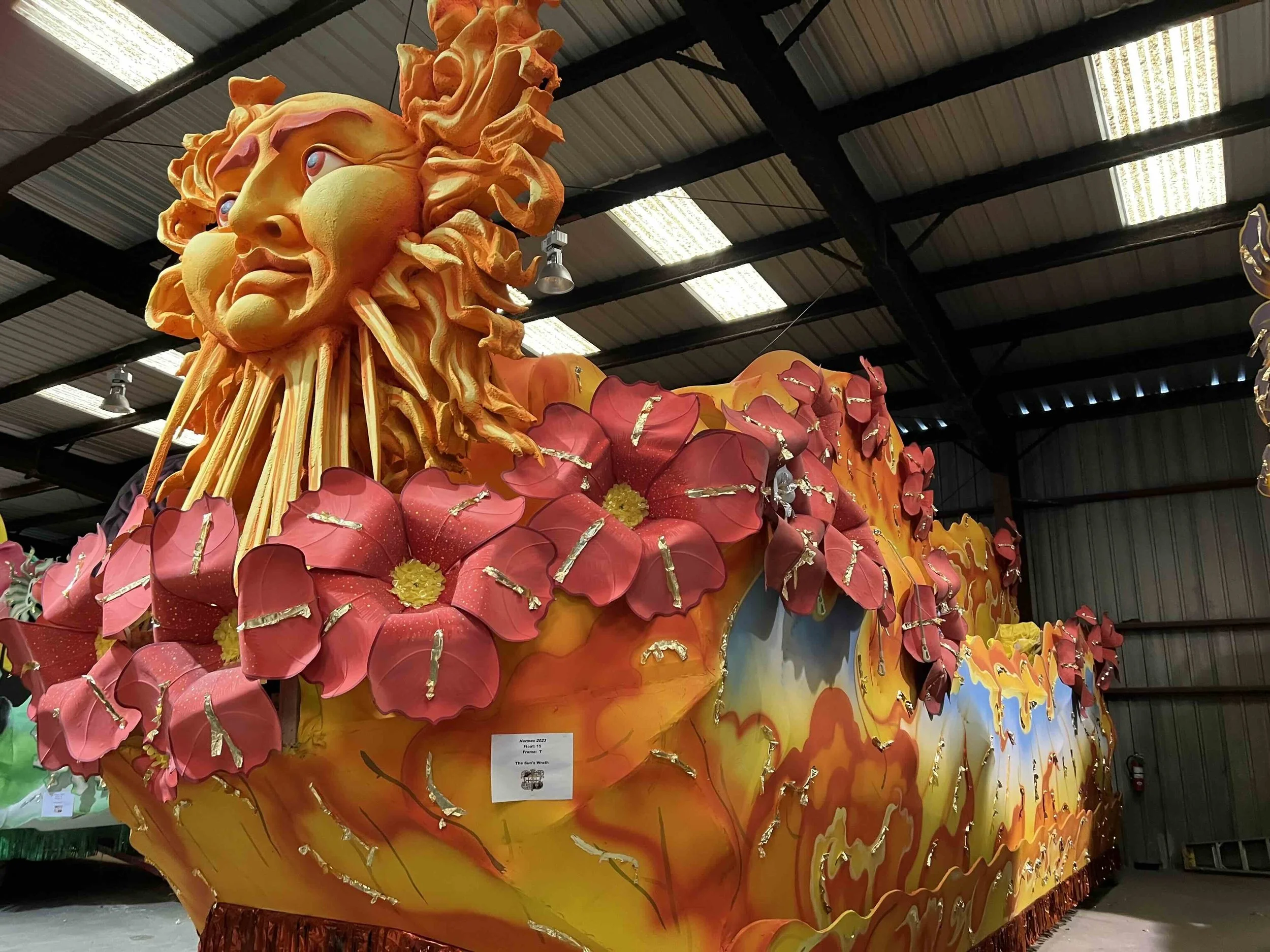 Large colorful parade float featuring a golden sun with a face and flowing rays, surrounded by red and pink flowers with gold accents, inside a warehouse.