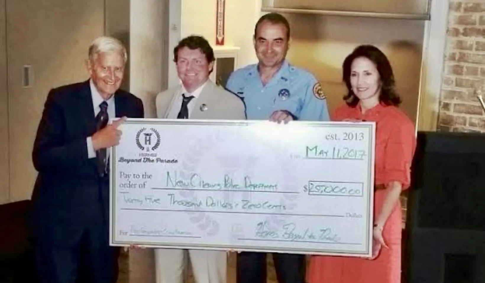 Five people holding a large ceremonial check. The check is made out to New Orleans Blue Dreams for $50,000 and is dated May 11, 2018. The group includes a man in a suit, a man in a white uniform, a man in a police uniform, and a woman in a red dress, standing indoors with a brick wall and a painting in the background.