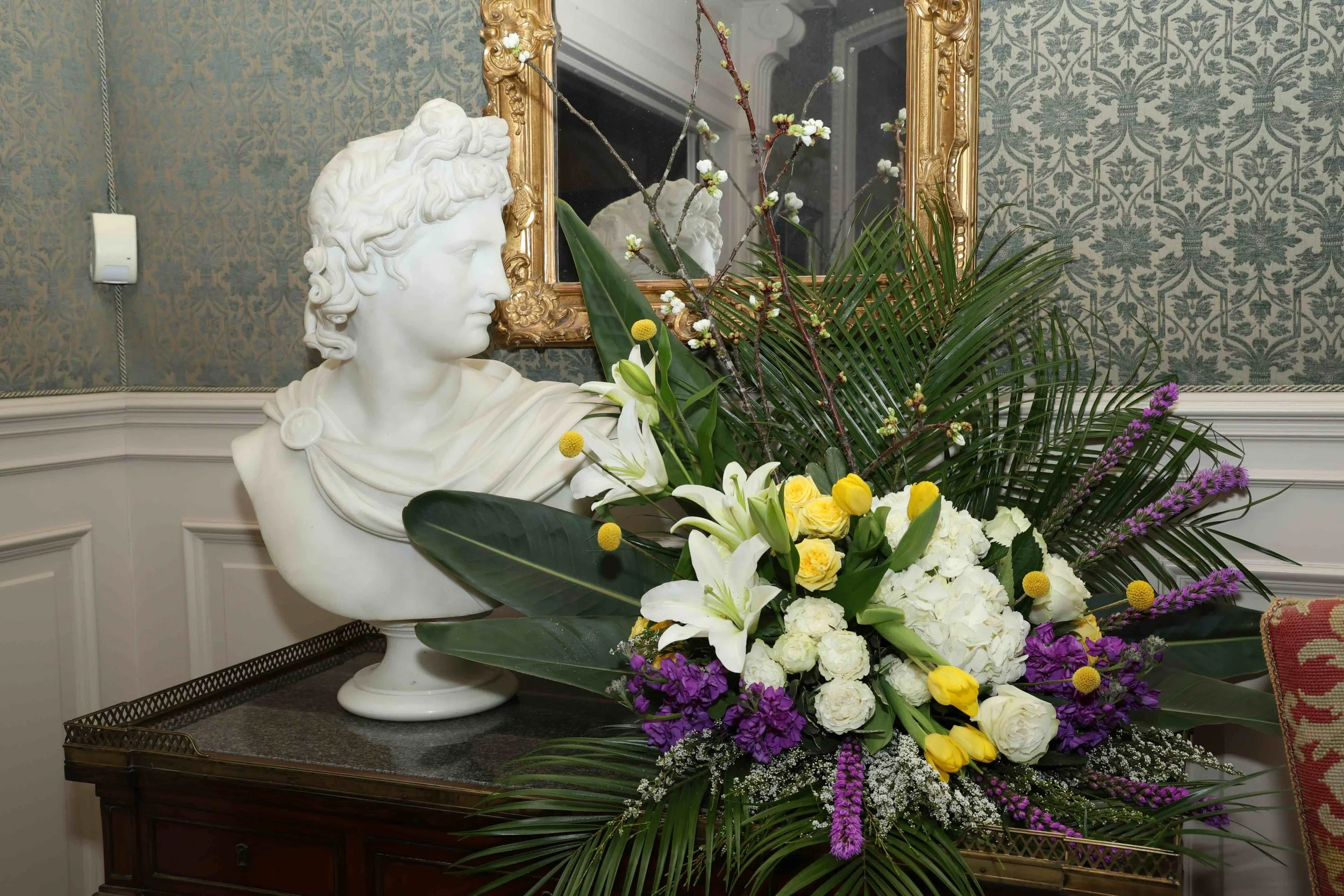 White marble bust of a classical man with curly hair and a cloak, surrounded by a vibrant bouquet of yellow, white, purple, and green flowers and leaves, set on a dark wooden table in a room with ornate wallpaper and a gold-framed mirror in the background.
