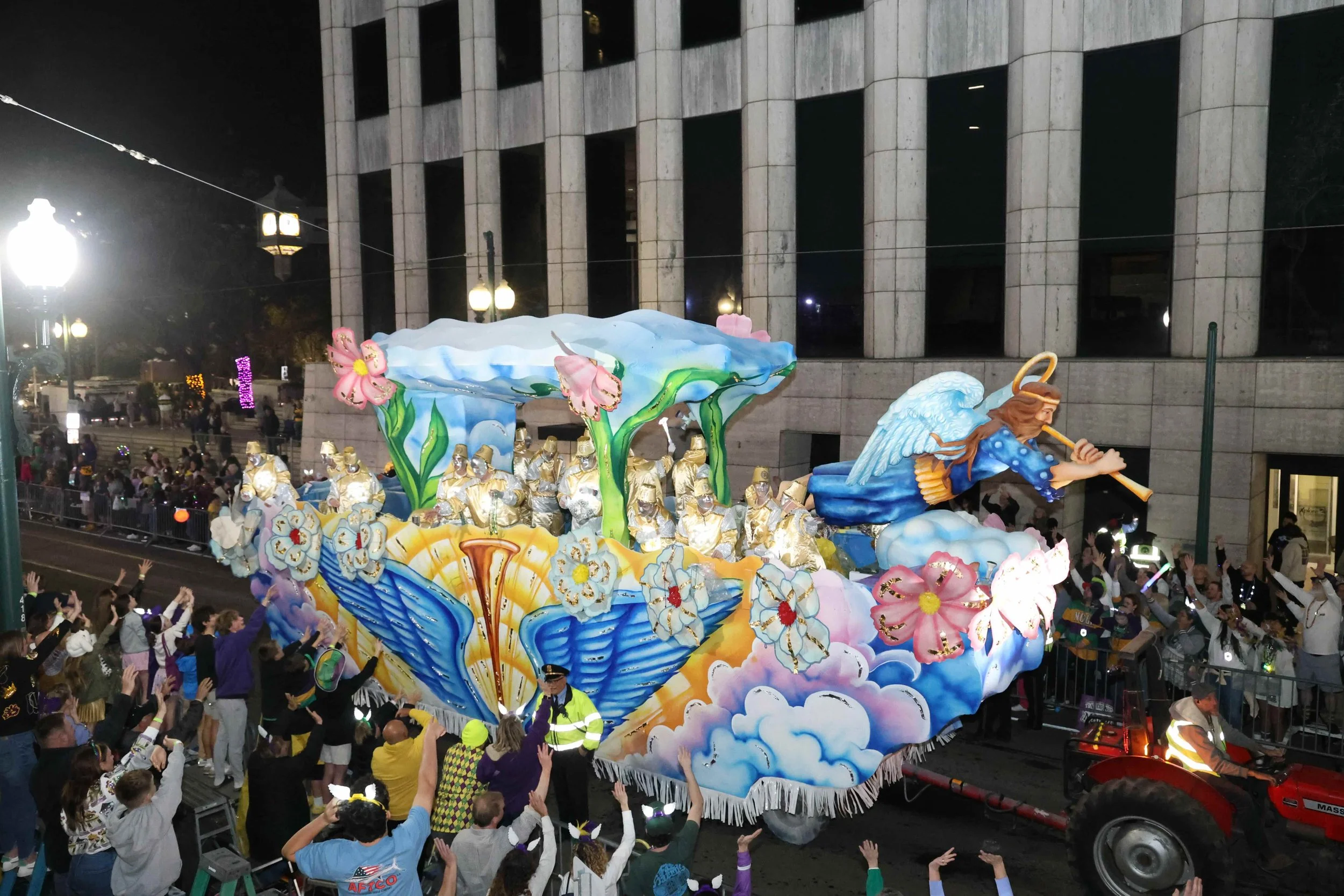 A colorful Hermes parade float decorated with flowers, clouds, and a large angel playing a flute, surrounded by performers and spectators at night.