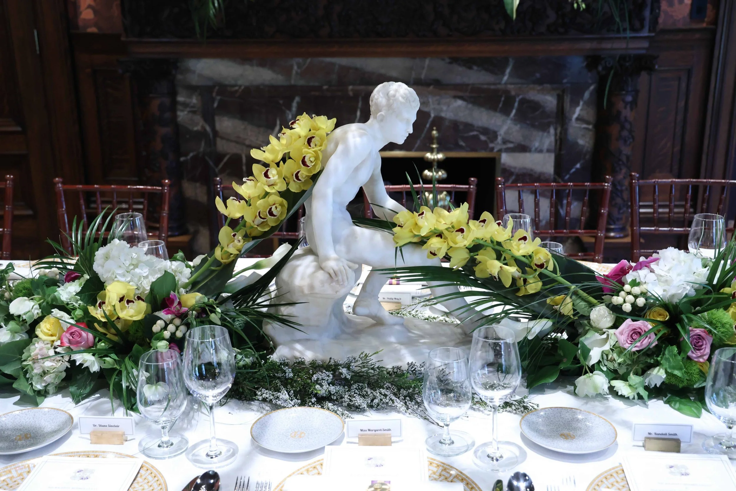 A table decorated with floral arrangements and a white marble statue of Hermes, surrounded by yellow orchids, white, pink, and purple roses, and greenery, with empty wine glasses and plates, in a formal dining setting.