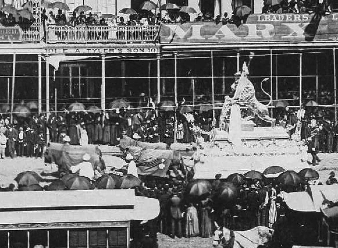 Historical black-and-white photograph of a crowded street scene with a large crowd of people and an elaborate statue or monument in the center, surrounded by ornamental fencing. People are holding umbrellas, possibly indicating rain, and some are walking or standing near a building with signs, including
