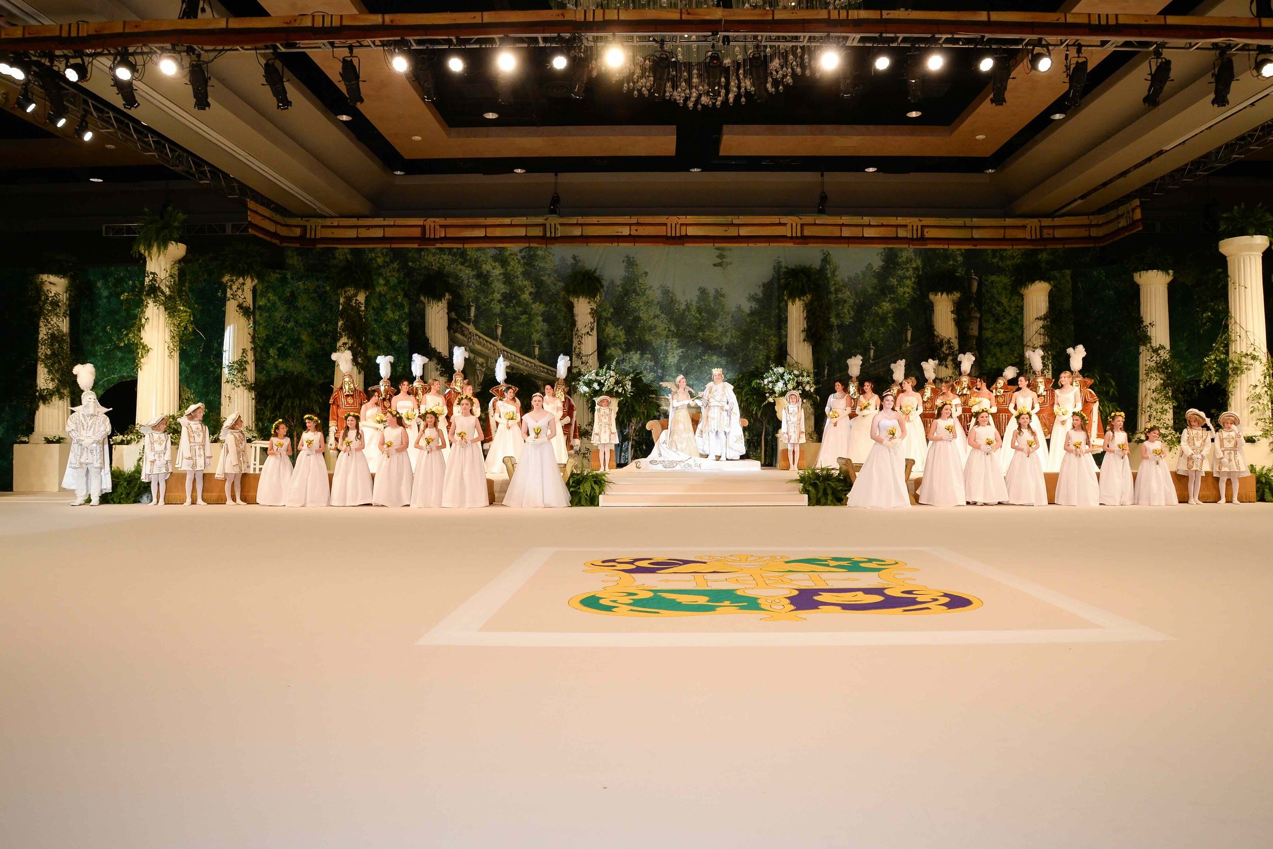 A large theatrical stage set to resemble an ancient Greek garden with columns, greenery, and a backdrop of trees. The stage features two central figures, likely a bride and groom, dressed in elaborate costumes, with a group of people dressed in period costumes and white dresses surrounding them, forming a ceremonial or celebratory scene. The foreground has a decorated floor with a crest or emblem.