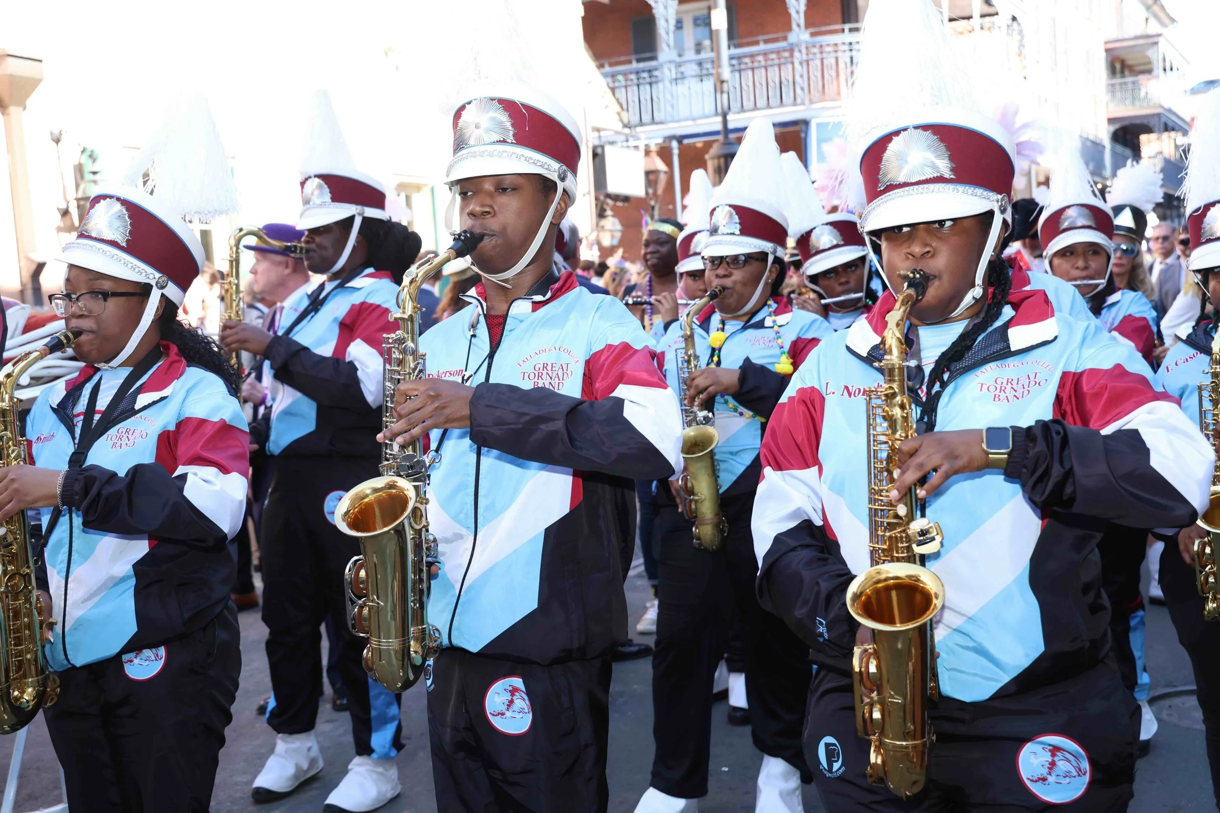 A marching band with saxophones, dressed in band uniforms with light blue, black, and red colors, and wearing white hats with feathers.