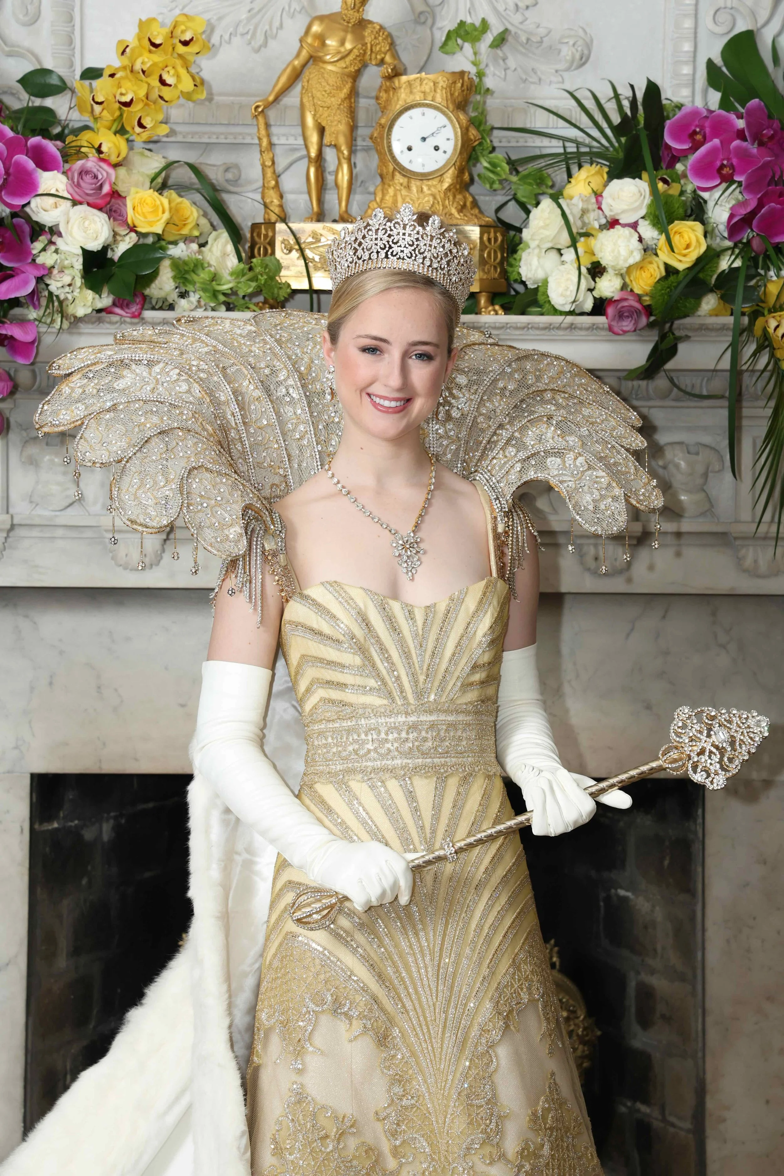 A woman dressed as a queen or princess, wearing a gold and cream gown with ornate shoulder embellishments, a tiara, and white gloves, holding a regal scepter, standing in front of a fireplace decorated with colorful flowers and a clock.