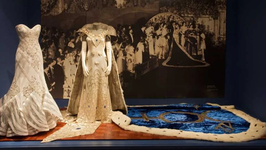 Display of two vintage wedding dresses and a large, ornate wedding train in a museum exhibit, with a black-and-white wedding scene mural in the background.