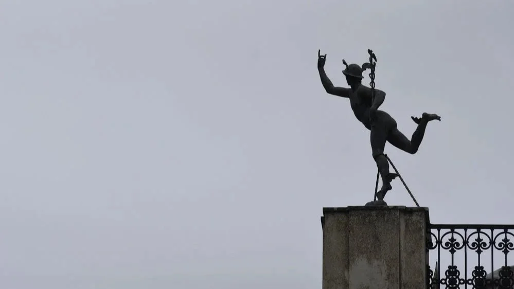Silhouette of a statue of the Greek God Hermes, standing on one leg on a ledge, with an ornate railing nearby and a cloudy sky background.