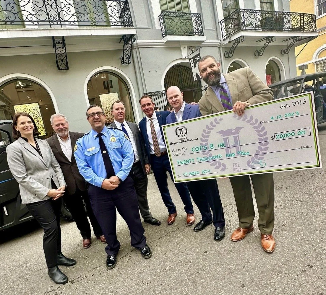 A group of five men and one woman standing outside a building, holding a large ceremonial check for $20,000 made out to COPs 8, Inc., dated December 4, 2023, from Hermes Beyond The Parade, established in 2013. The man on the right is holding the check and smiling.