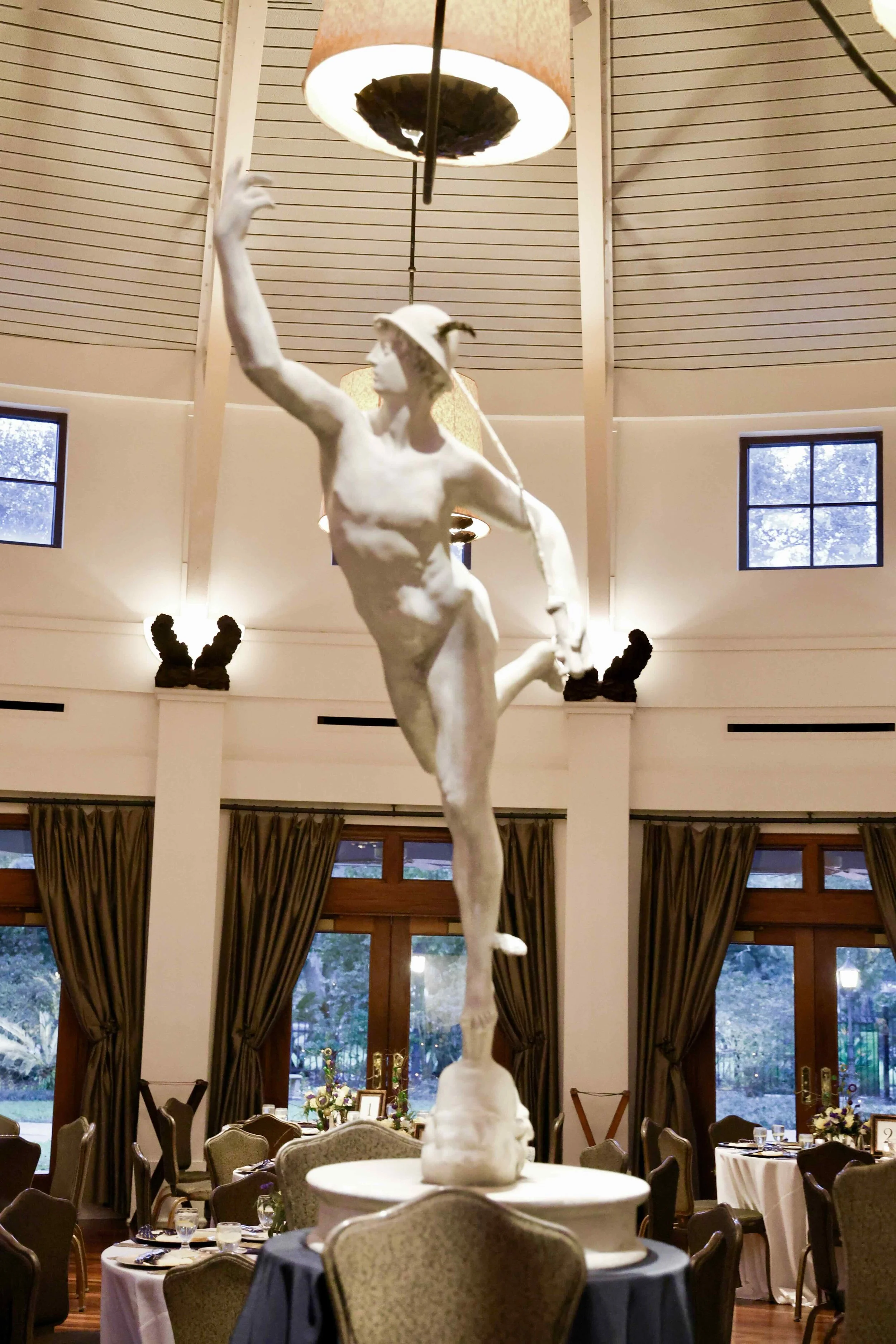 A large sculpture of the Greek God Hermes is displayed in a banquet hall with tables, chairs, and large windows.