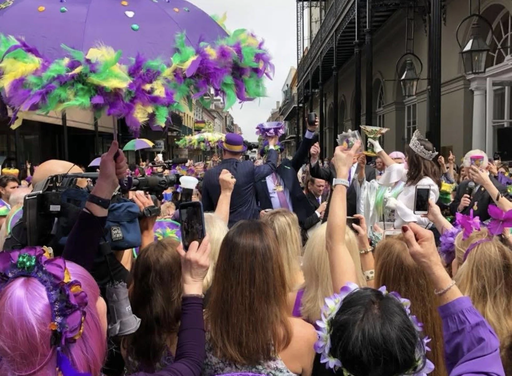 Crowd gathered in a street parade dressed in purple, green, and yellow, many wearing hats and crowns, with feathers and flowers, holding umbrellas and taking photos. The buildings have balcony railings and lanterns.