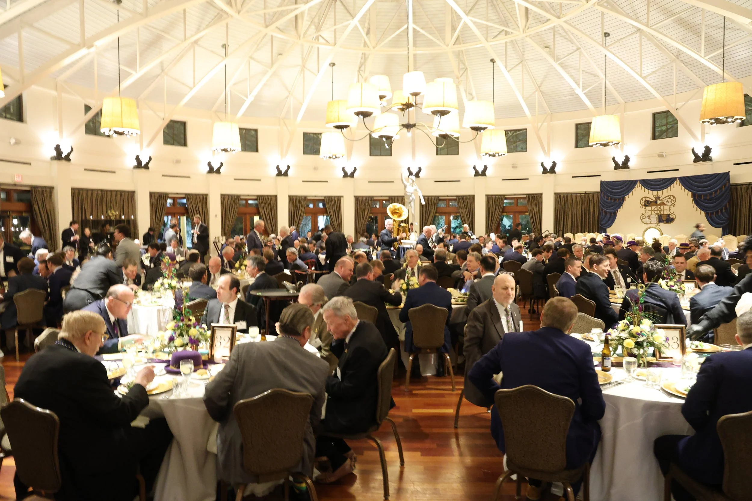 A large banquet hall filled with many round tables, where people in formal attire are seated for what appears to be a formal event or dinner, with high ceiling, chandeliers, and elegant drapery.