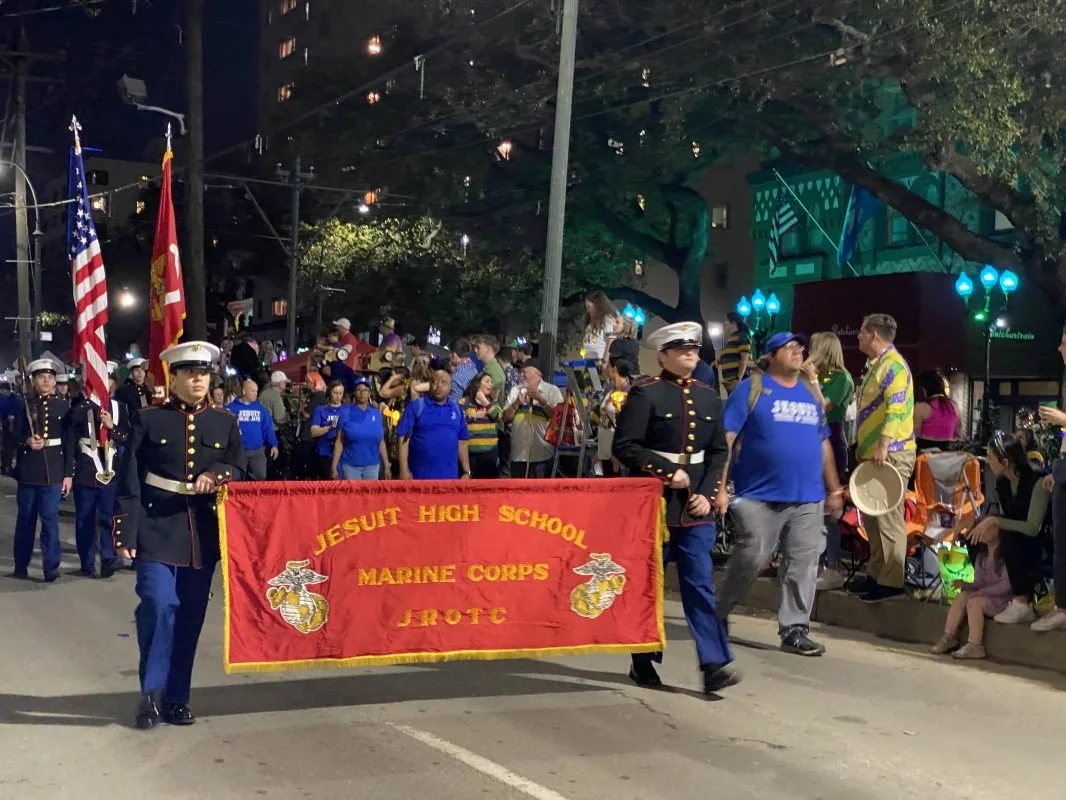 A parade with Jesuit High School Marine Corps JROTC students marching and carrying a red banner, with spectators watching along the street at night.