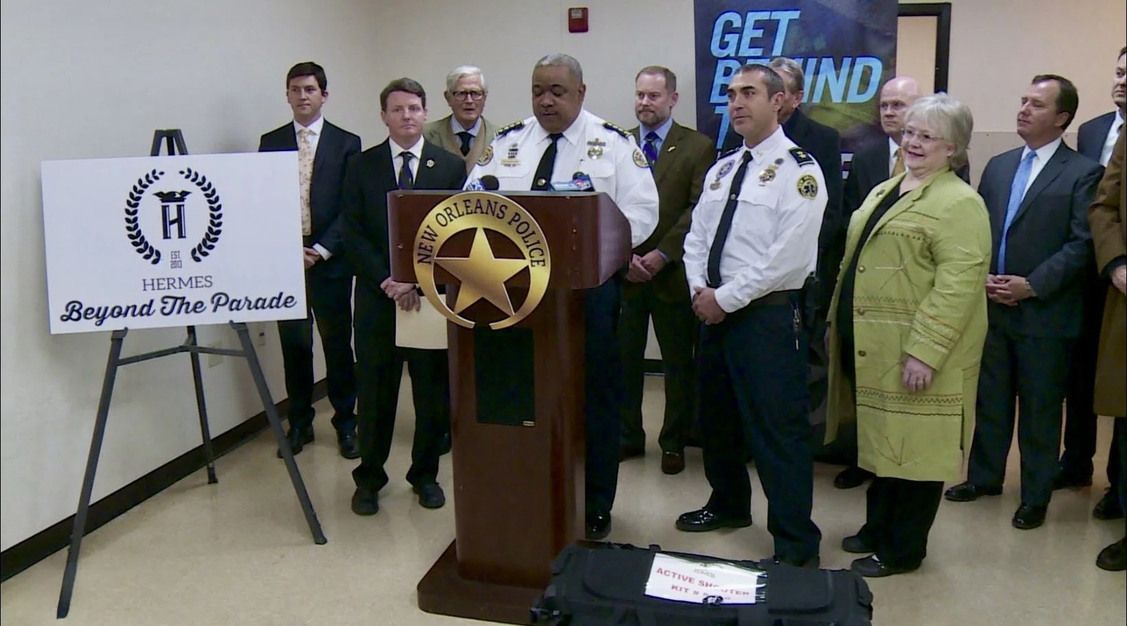 A group of people, including police officers and officials, standing behind a wooden podium with a New Orleans Police emblem, during a press conference. There is a sign on an easel to the left that reads "Hermes Beyond The Parade." In the background, a poster says "Get Back."