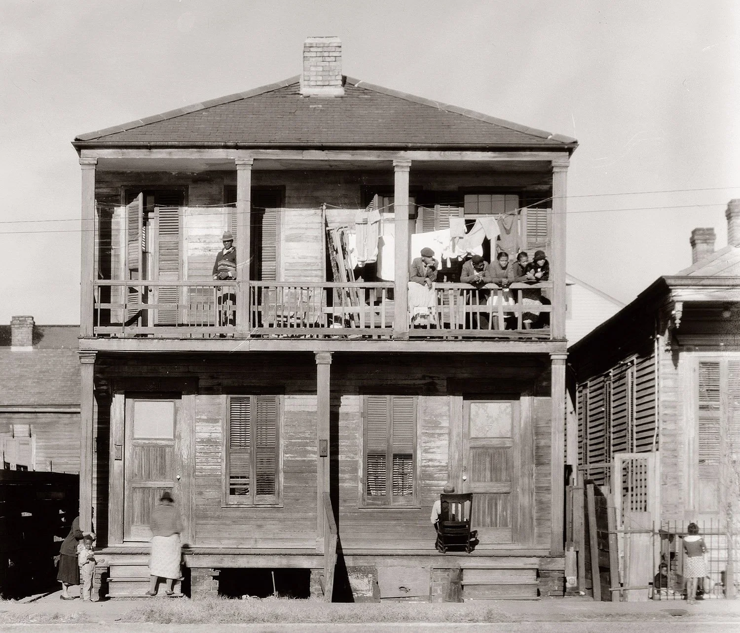 A two-story wooden house with a porch on both levels. People are sitting on the second-floor porch, and laundry is hanging on a line. A woman and children are standing at the front steps, and a man is standing on the second-floor porch among others.