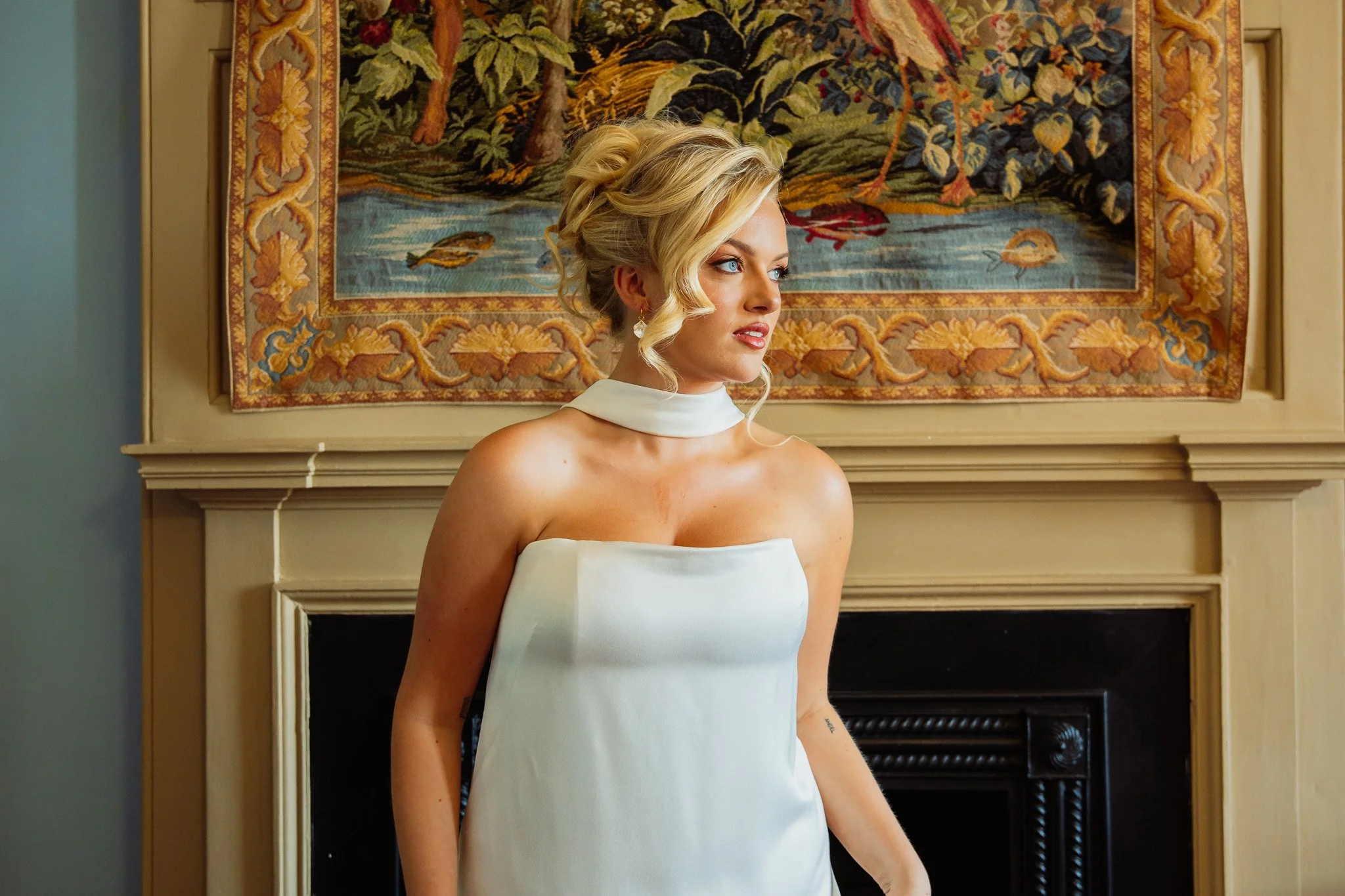 A woman with blonde hair in styled curls, wearing a white strapless dress with a white choker, standing in front of a detailed tapestry hanging above a fireplace.