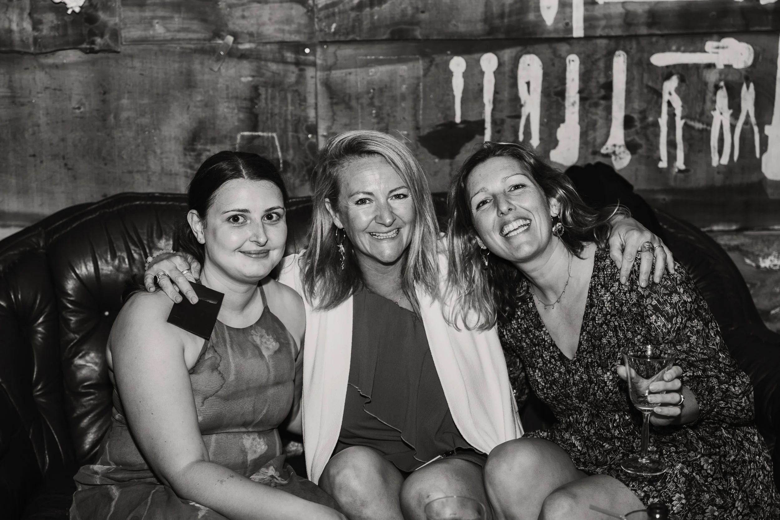 Three women sitting closely on a leather couch, smiling and embracing each other, in a dimly lit room with wooden walls and a sign in the background. One woman holds a glass of wine.