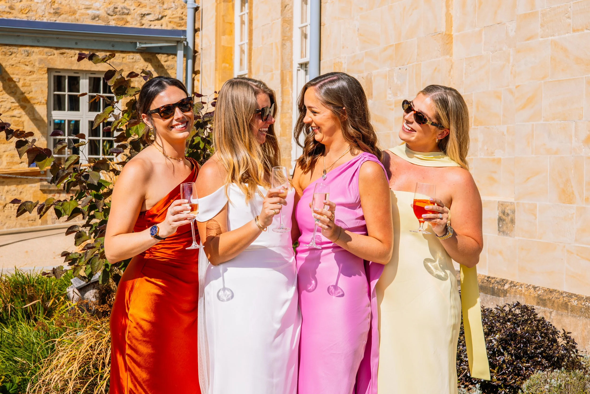 Four women in colorful dresses smiling and holding glasses of wine or champagne in an outdoor setting with a stone building and greenery.