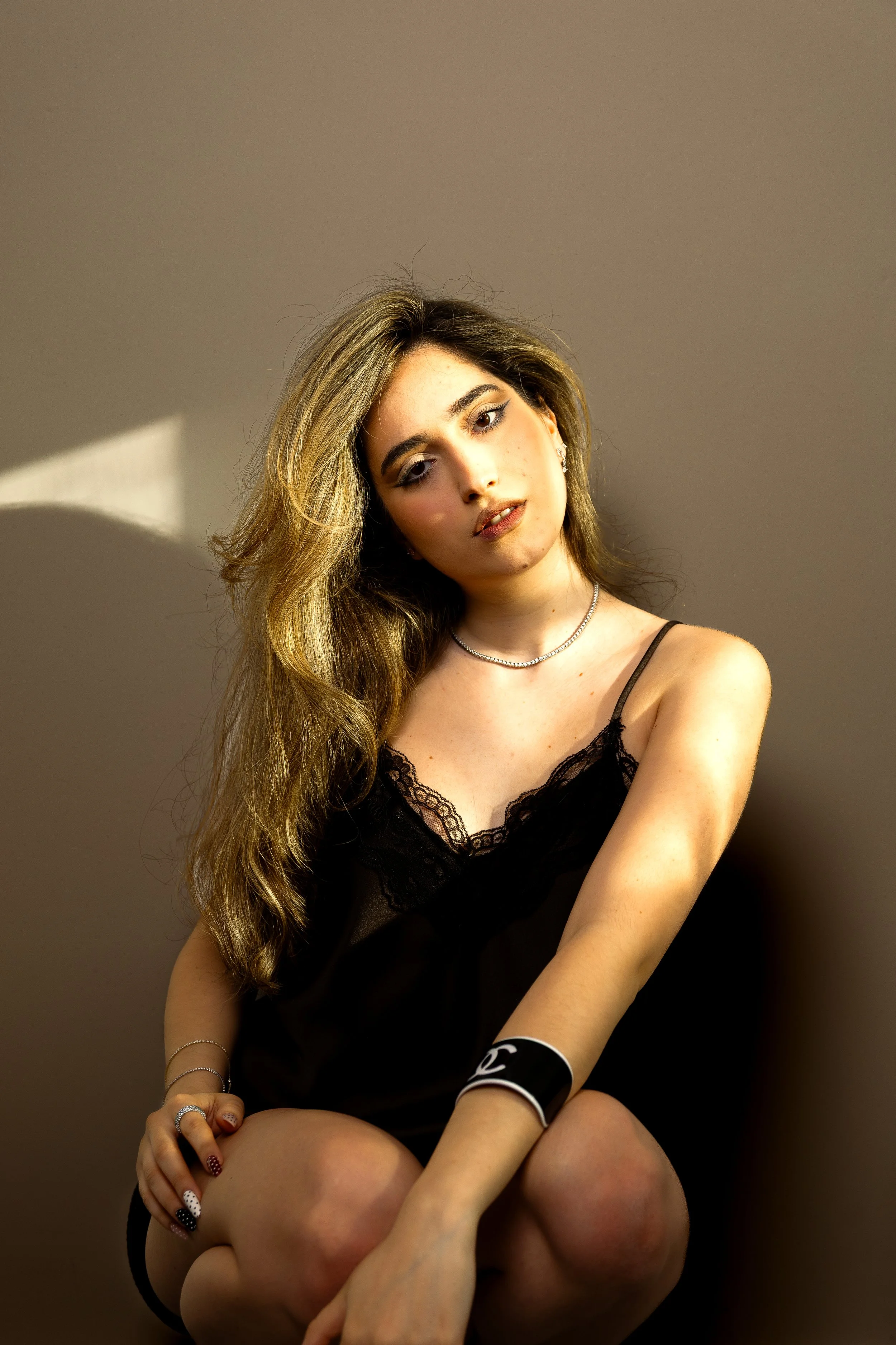 A young woman with long, wavy light brown hair, wearing a black lace camisole, jewelry, and makeup, poses against a plain wall with her head tilted and her arms wrapped around her knees.