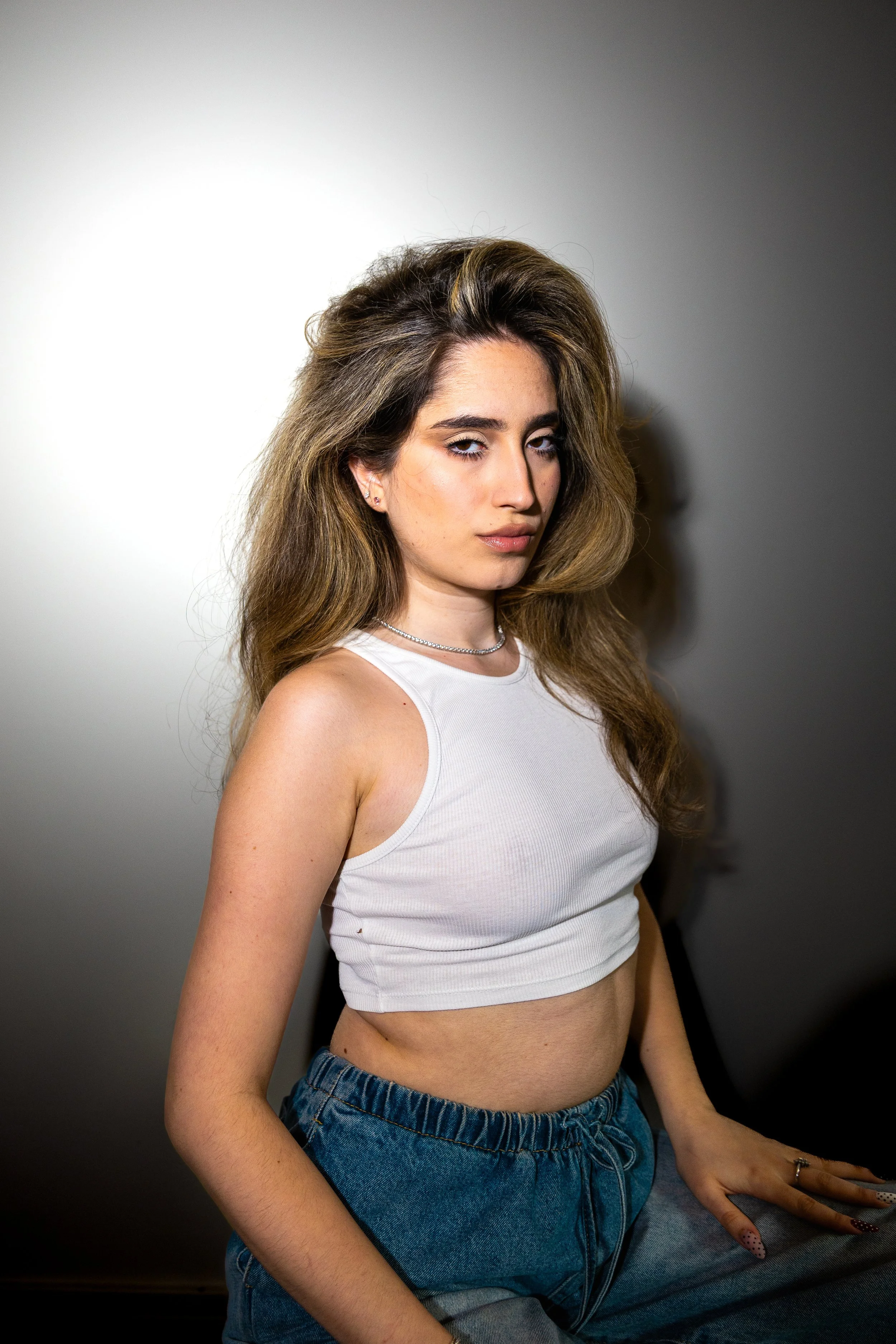 A young woman with long, wavy brown hair and light skin, wearing a white sleeveless crop top and blue denim jeans, posing against a gradient background with a shadow cast to her right.