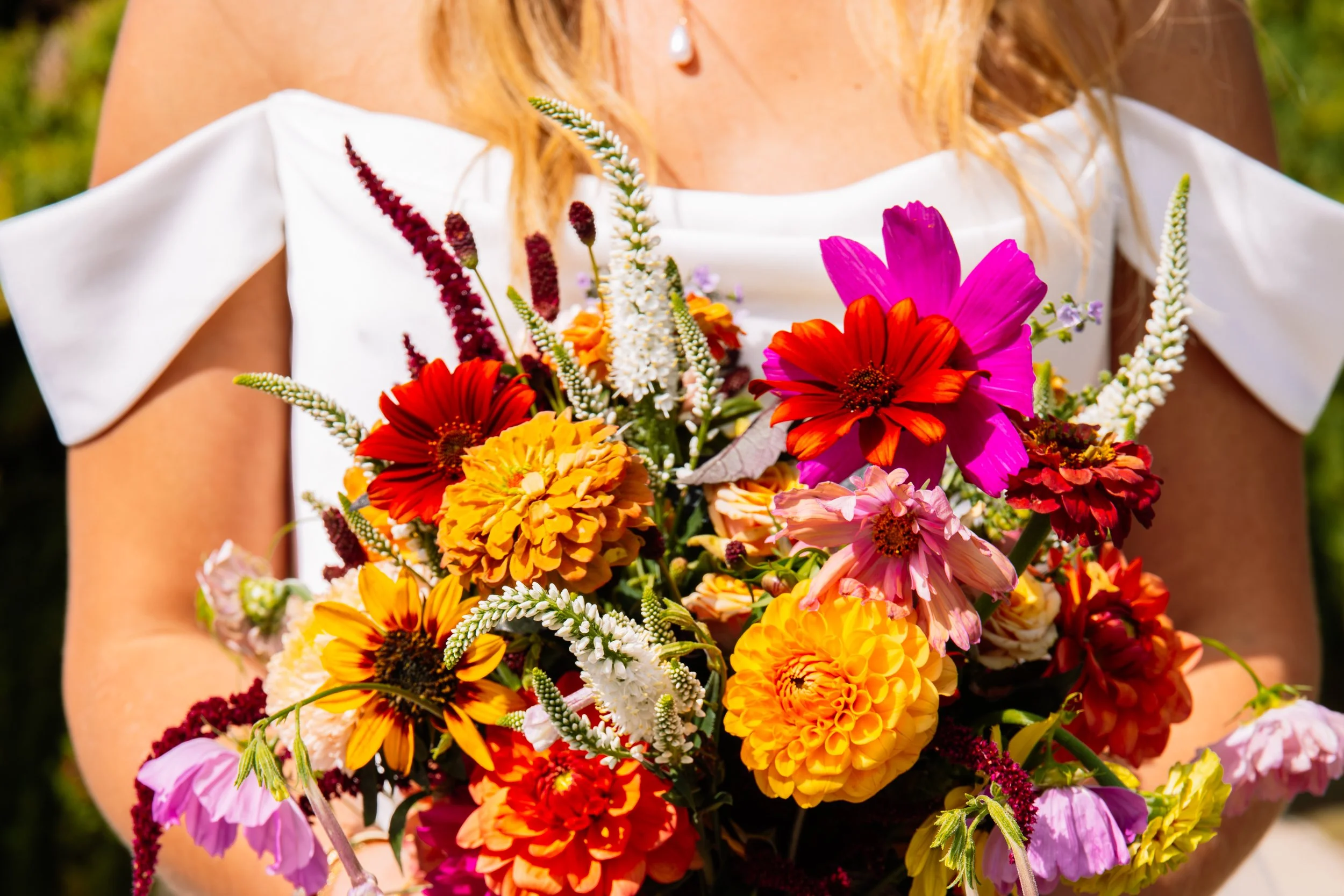 A woman holding a large bouquet of colorful flowers including pink, red, orange, yellow, and white blooms, with a blurred outdoor background.